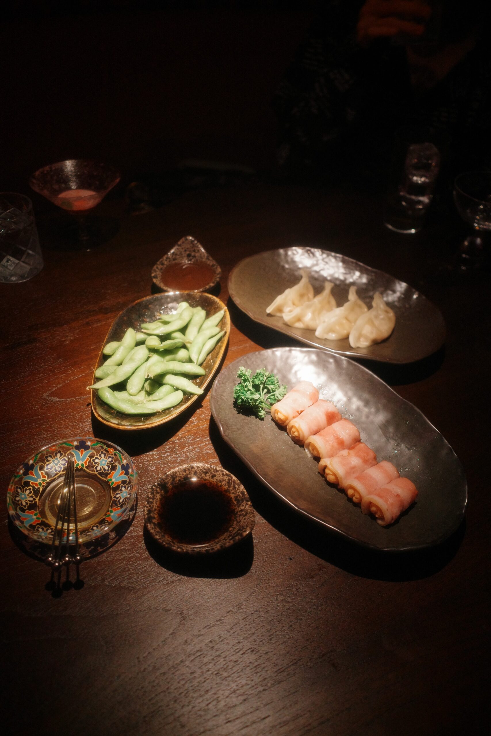 จานอาหารญี่ปุ่นสไตล์อิซากายะบนโต๊ะไม้ มีถั่วแระญี่ปุ่น เกี๊ยวซ่านึ่ง เบคอนม้วน พร้อมซอสจิ้มและเครื่องดื่ม ในบรรยากาศร้านแสงสลัว