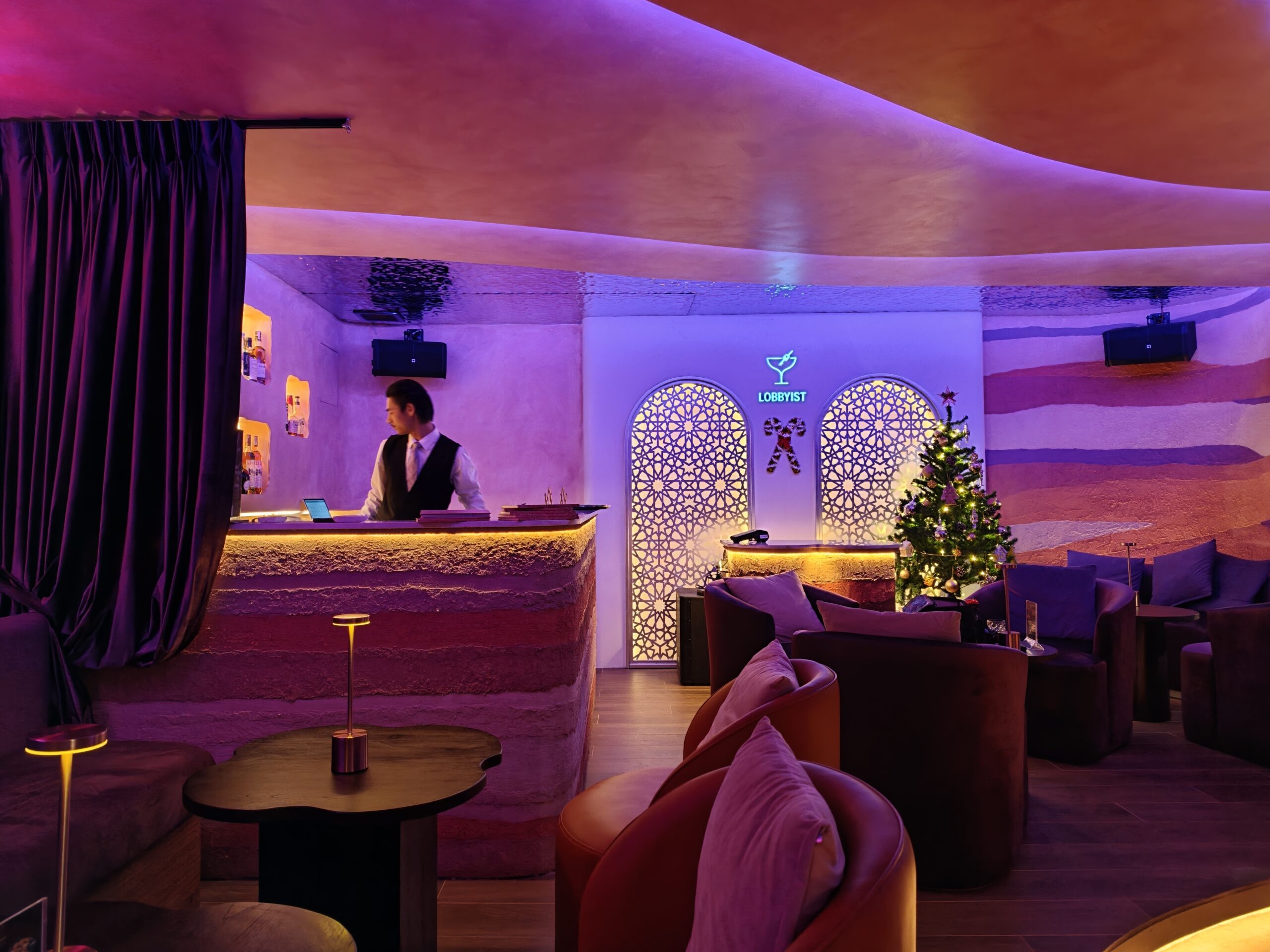 บาร์ Lobbyist ตกแต่งสไตล์โมร็อกโกด้วยแสงสีม่วง มีบาร์เทนเดอร์ยืนหลังเคาน์เตอร์ โซฟานุ่มสบาย และต้นคริสต์มาสประดับไฟ