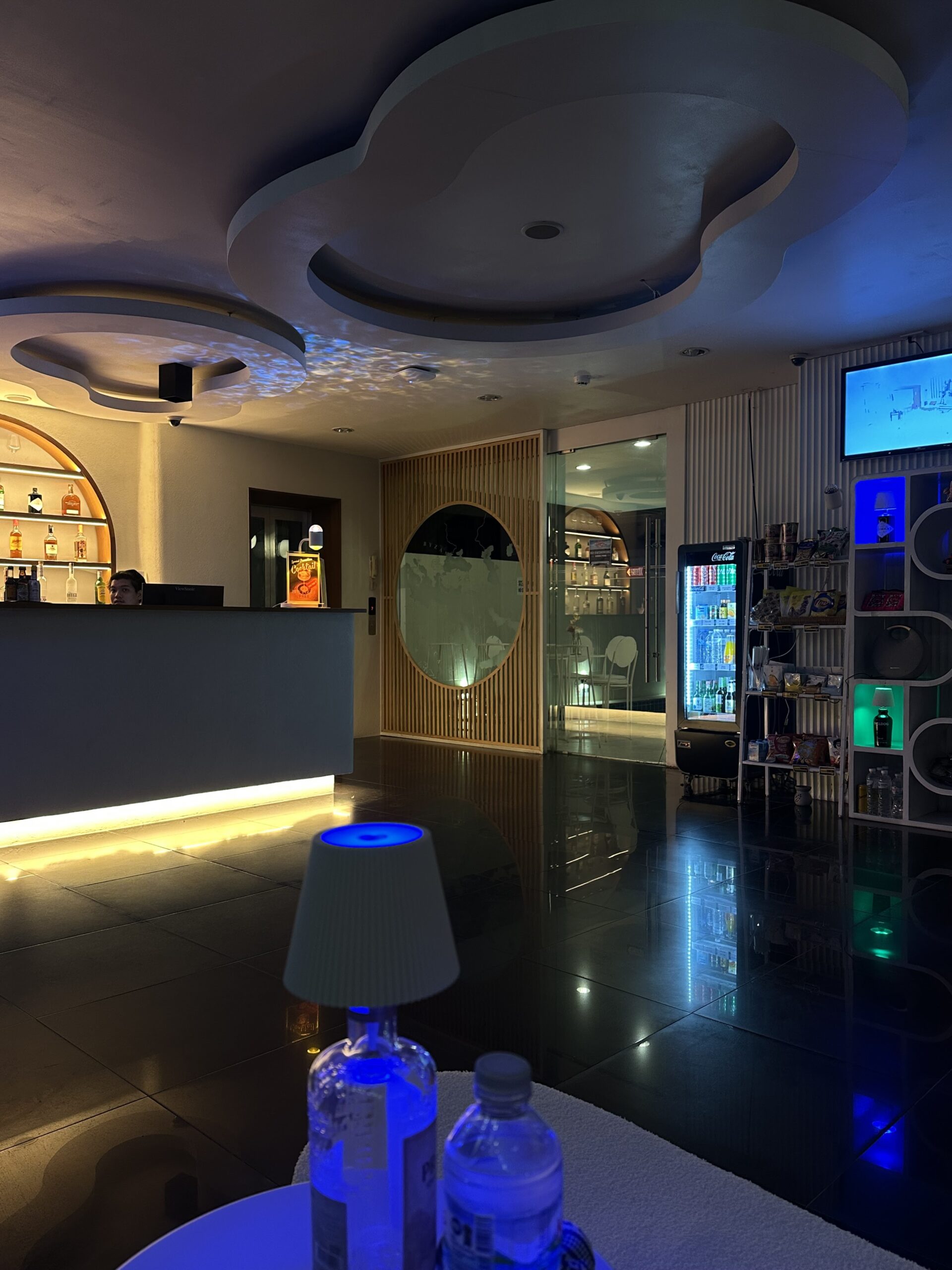 บาร์เลานจ์สไตล์โมเดิร์นพร้อมไฟบรรยากาศสีน้ำเงิน ตู้เครื่องดื่มเย็น ชั้นวางเหล้า และโคมไฟตั้งโต๊ะ