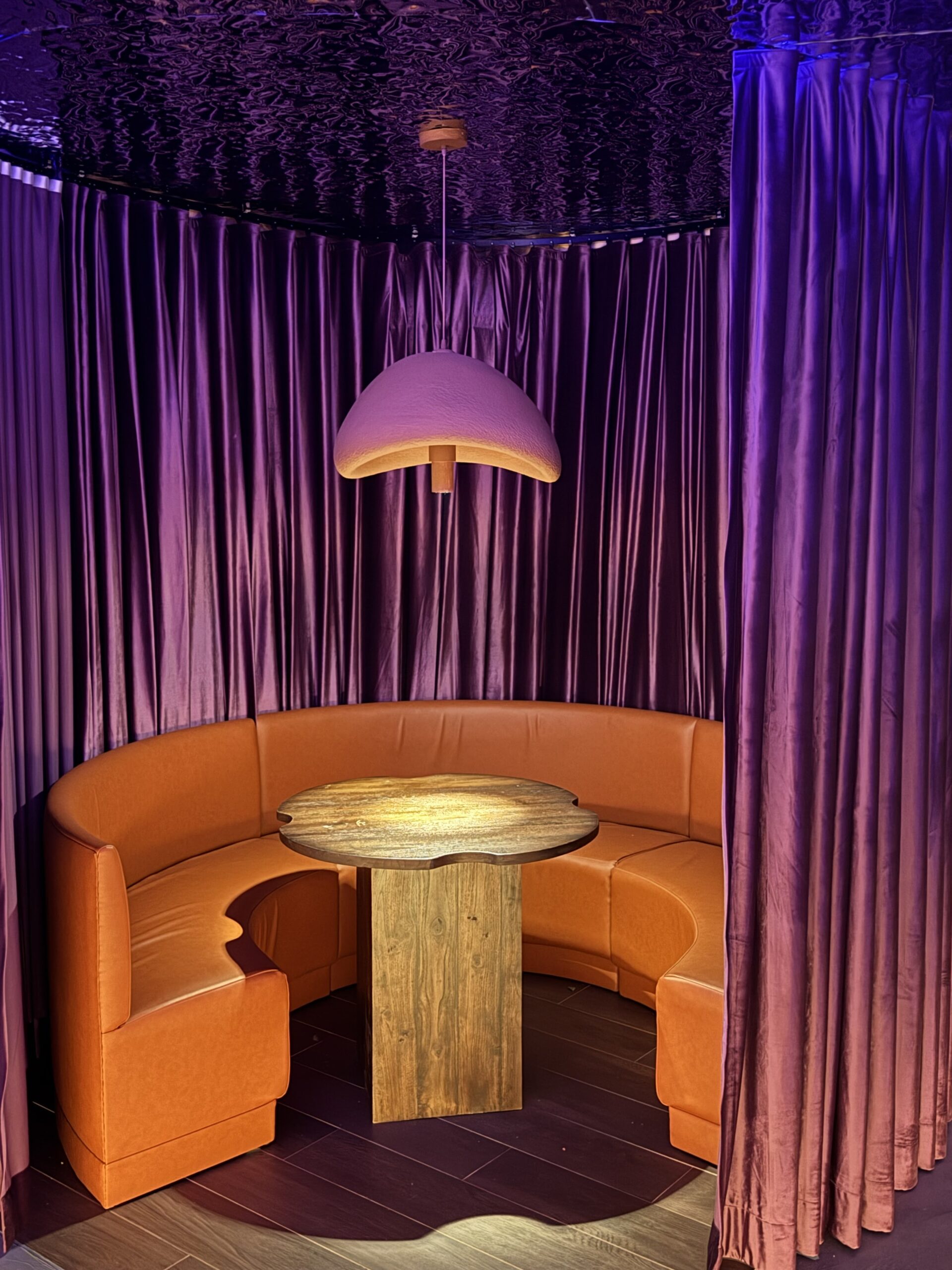 มุมนั่งบูธหนังสีส้มรูปโค้งพร้อมโต๊ะไม้และโคมไฟแขวนในห้องผ้าม่านกำมะหยี่สีม่วง