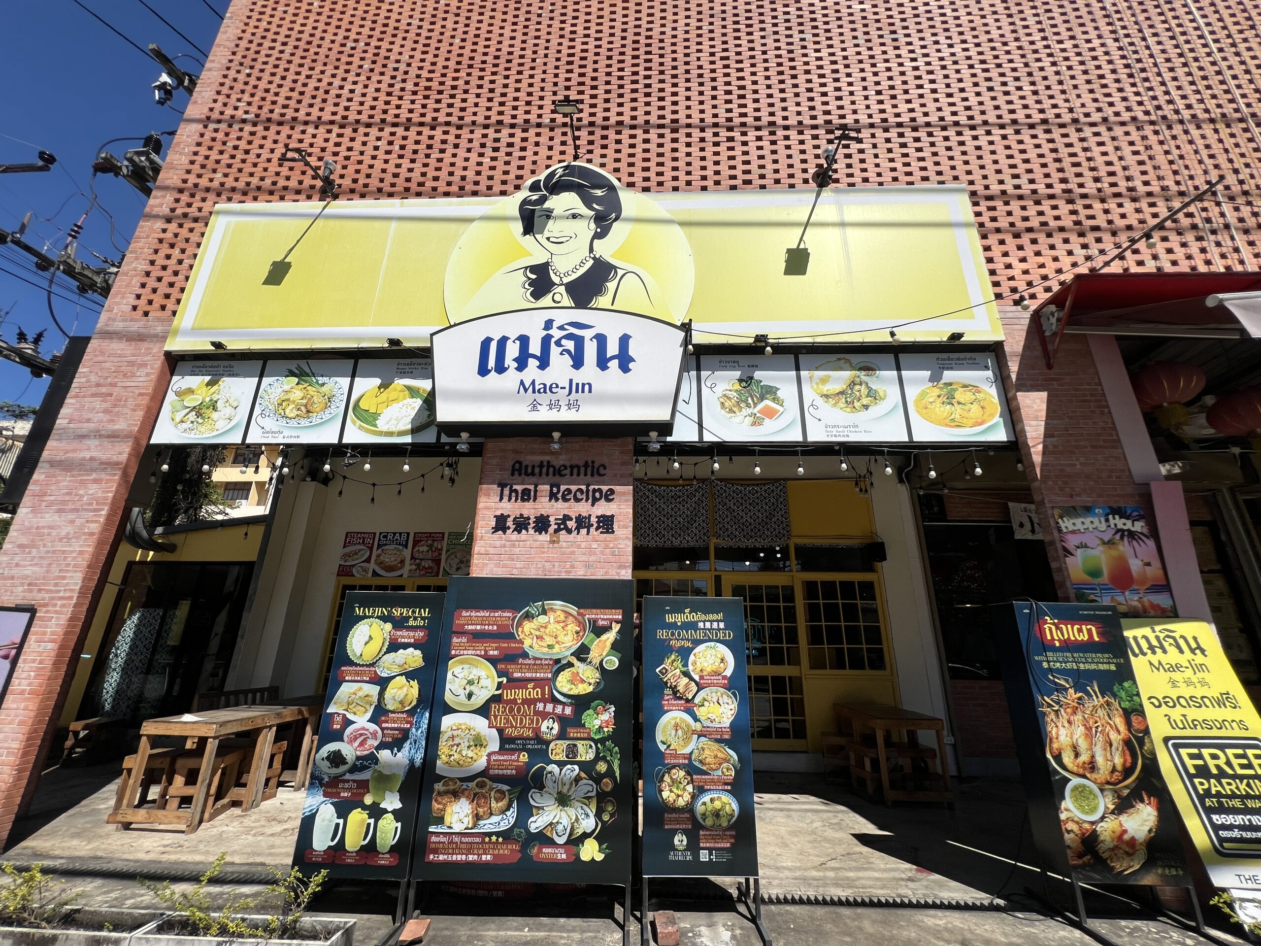 ร้านอาหารแม่จิน หน้าร้านอาหารไทยแท้ มีป้ายเมนูแนะนำและภาพอาหารตั้งหน้าร้าน อาคารอิฐแดง ป้ายสีเหลืองรูปคุณแม่จิน พร้อมข้อความ Authentic Thai Recipe