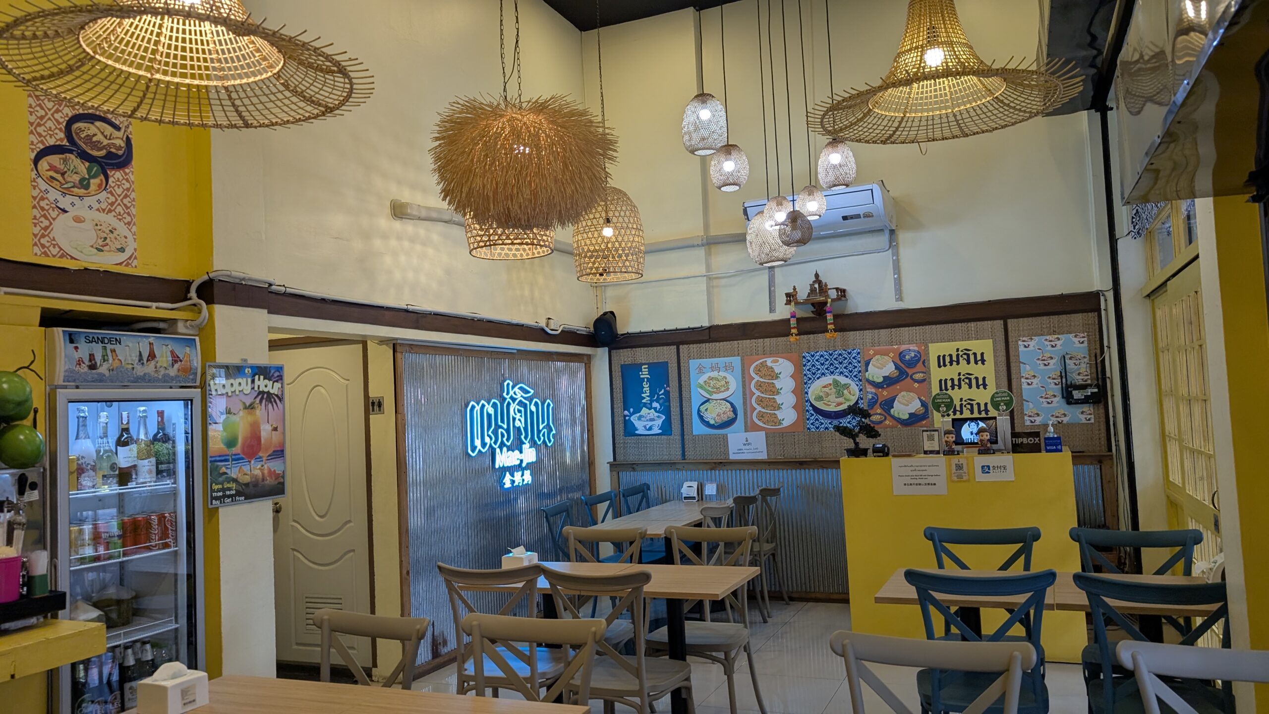 ภายในร้านแม่จิน ตกแต่งด้วยโคมไฟหวายทรงหมวกและโทนสีเหลือง มีโต๊ะเก้าอี้ไม้พร้อมป้ายเมนูอาหารติดผนัง