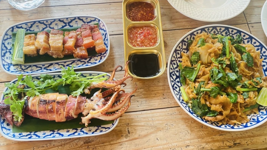 อาหารไทยบนจานลายครามสีน้ำเงิน มีหมูกรอบหั่นชิ้น ปลาหมึกย่าง ผัดซีอิ๊วใส่คะน้า พร้อมน้ำจิ้ม 3 ชนิดในถาดทองเหลือง บนโต๊ะไม้