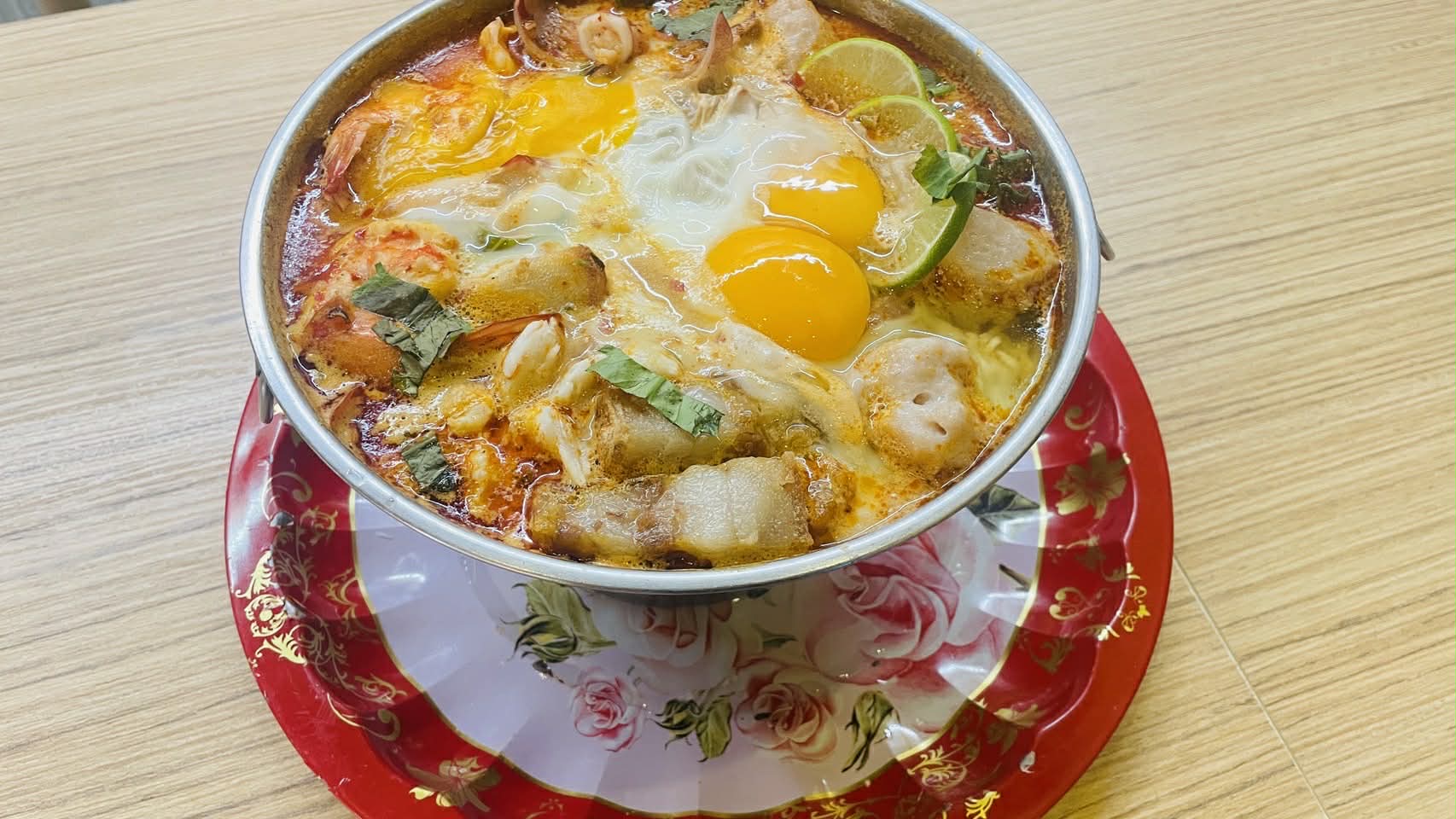 ต้มยำทะเลใส่ไข่ดาวเสิร์ฟในหม้อสแตนเลสบนจานลายดอกกุหลาบ มีกุ้ง หมึก หมูกรอบ มะนาว และผักชี