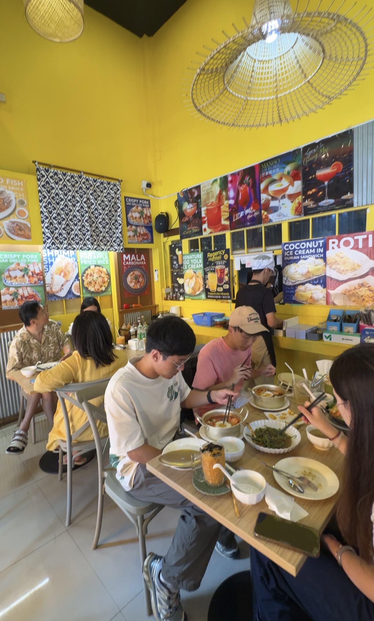 ร้านอาหารผนังสีเหลืองสดใสกับลูกค้าหลายกลุ่มนั่งทานอาหารไทยหลากหลายเมนู มีป้ายเมนูติดเต็มผนัง เช่น หมูกรอบ ข้าวผัดสับปะรด มะละกอคาโบนาร่า และไอศกรีมมะพร้าวในขนมปังทุเรียน