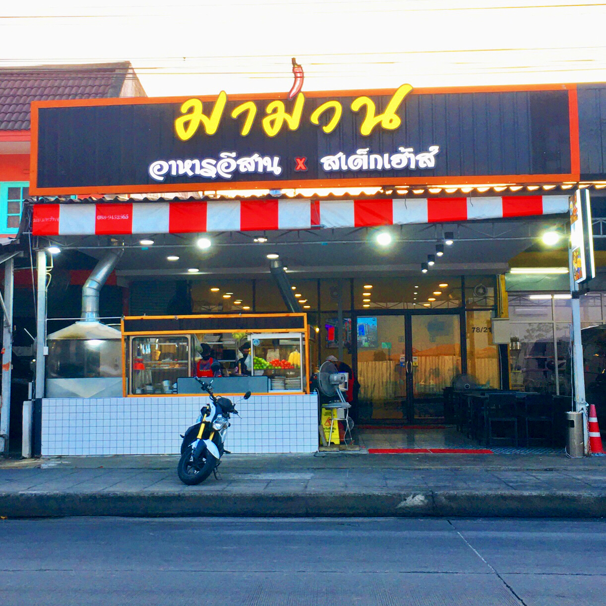 ร้านมามวน อาหารอีสาน x สเต็กเฮ้าส์ หน้าร้านพร้อมป้ายไฟสีเหลืองบนพื้นดำ มีหลังคาผ้าลายขาวแดง และมอเตอร์ไซค์จอดหน้าร้าน