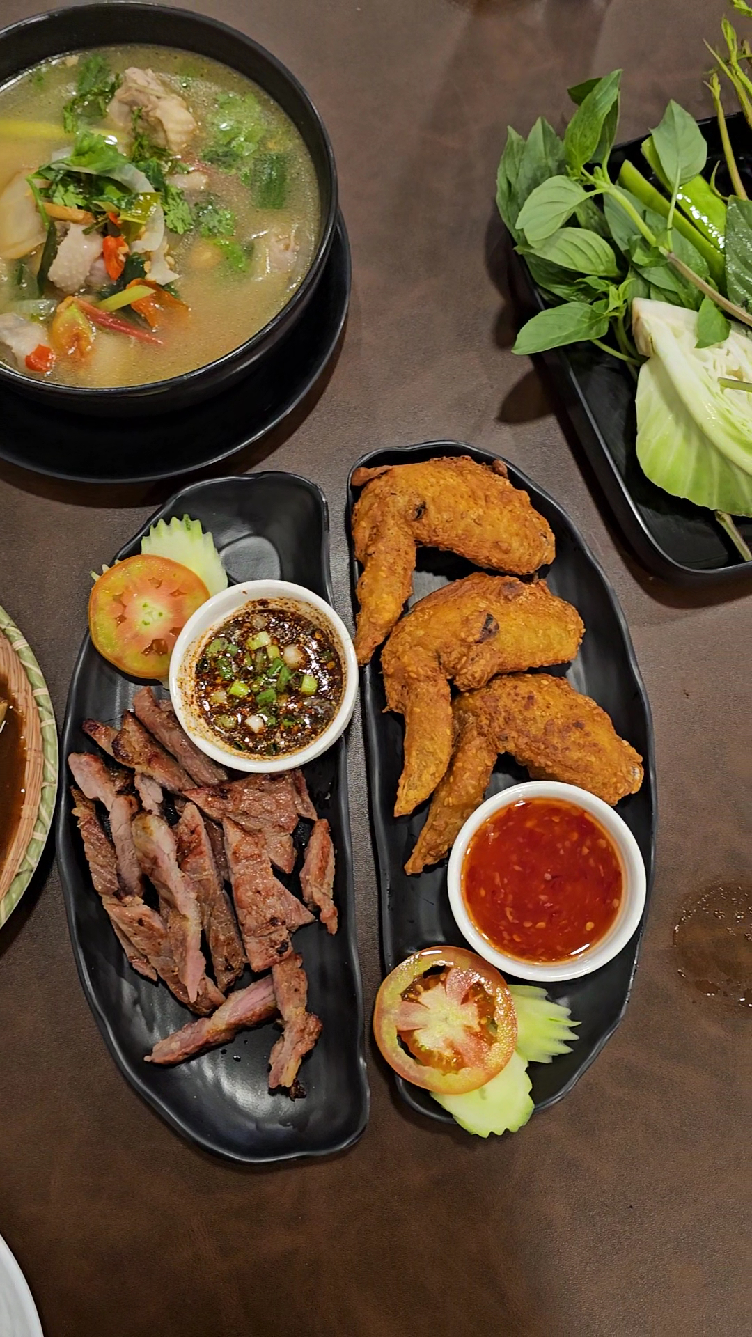 อาหารไทยบนโต๊ะ ประกอบด้วยเนื้อย่างหั่นชิ้นพร้อมน้ำจิ้มแจ่ว ปีกไก่ทอดพร้อมซอสพริก ต้มแซ่บ และผักสดเคียง