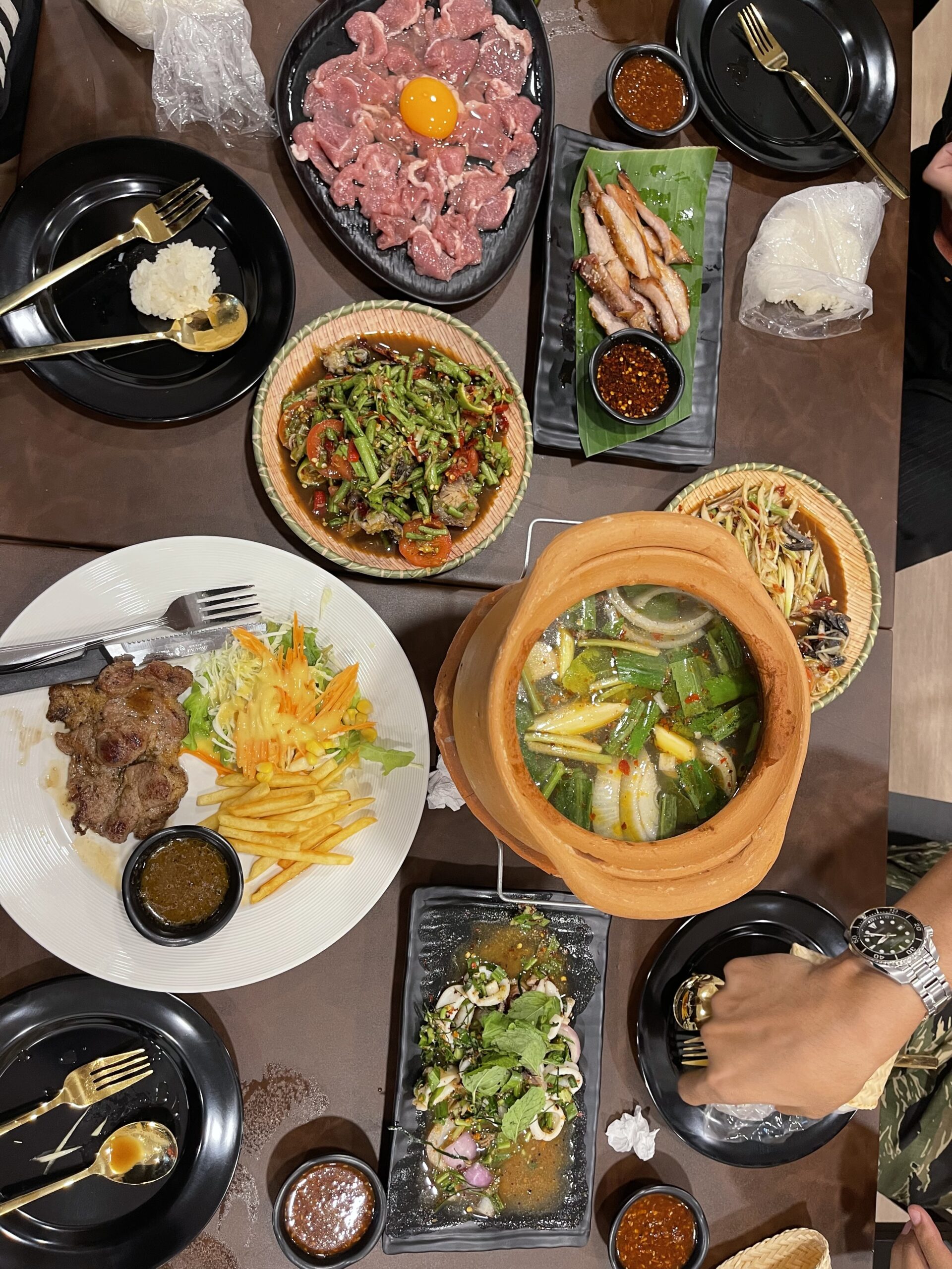 โต๊ะอาหารมองจากด้านบน มีเนื้อดิบพร้อมไข่แดง คอหมูย่าง ส้มตำ แกงร้อนในหม้อดิน สเต็กเนื้อกับเฟรนช์ฟรายส์ ยำทะเล และผัดพริกแดง พร้อมน้ำจิ้มหลายถ้วย