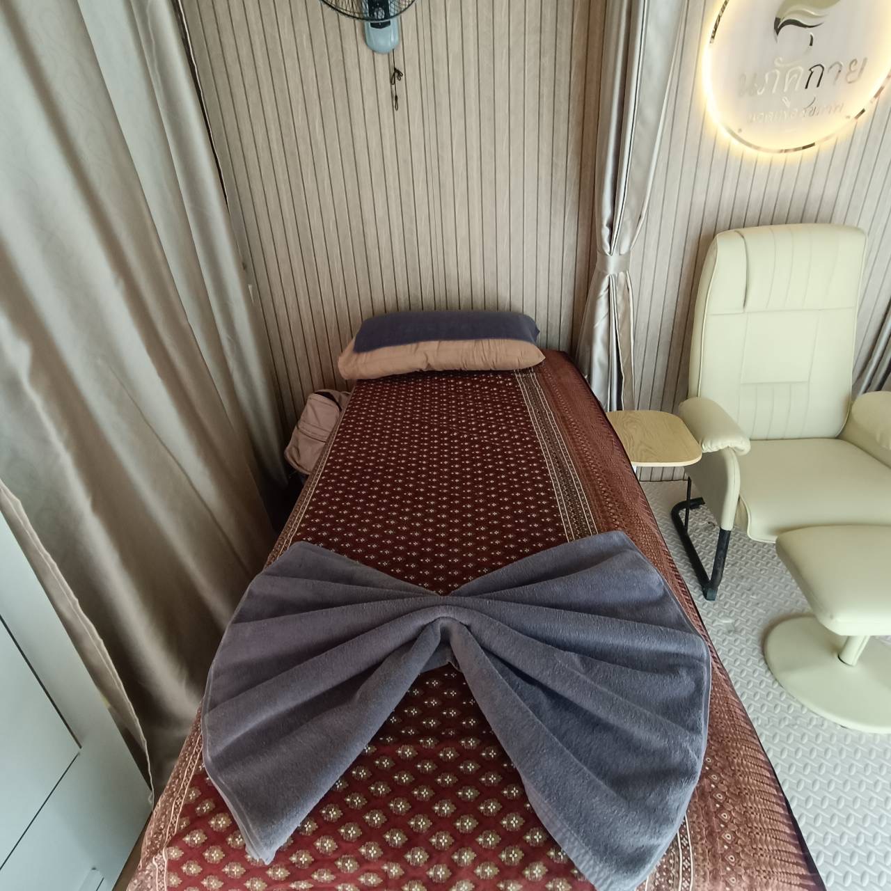 ห้องนวดไทยพร้อมเตียงนวดปูผ้าลายไทยสีแดงเลือดหมู ผ้าขนหนูสีเทาพับเป็นรูปโบว์ และเก้าอี้นวดสีขาว