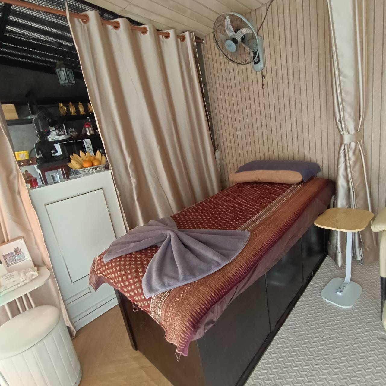 ห้องนวดไทยขนาดเล็กพร้อมเตียงนวดคลุมผ้าลายไทยสีแดง มีพัดลมติดผนังและม่านสีครีม