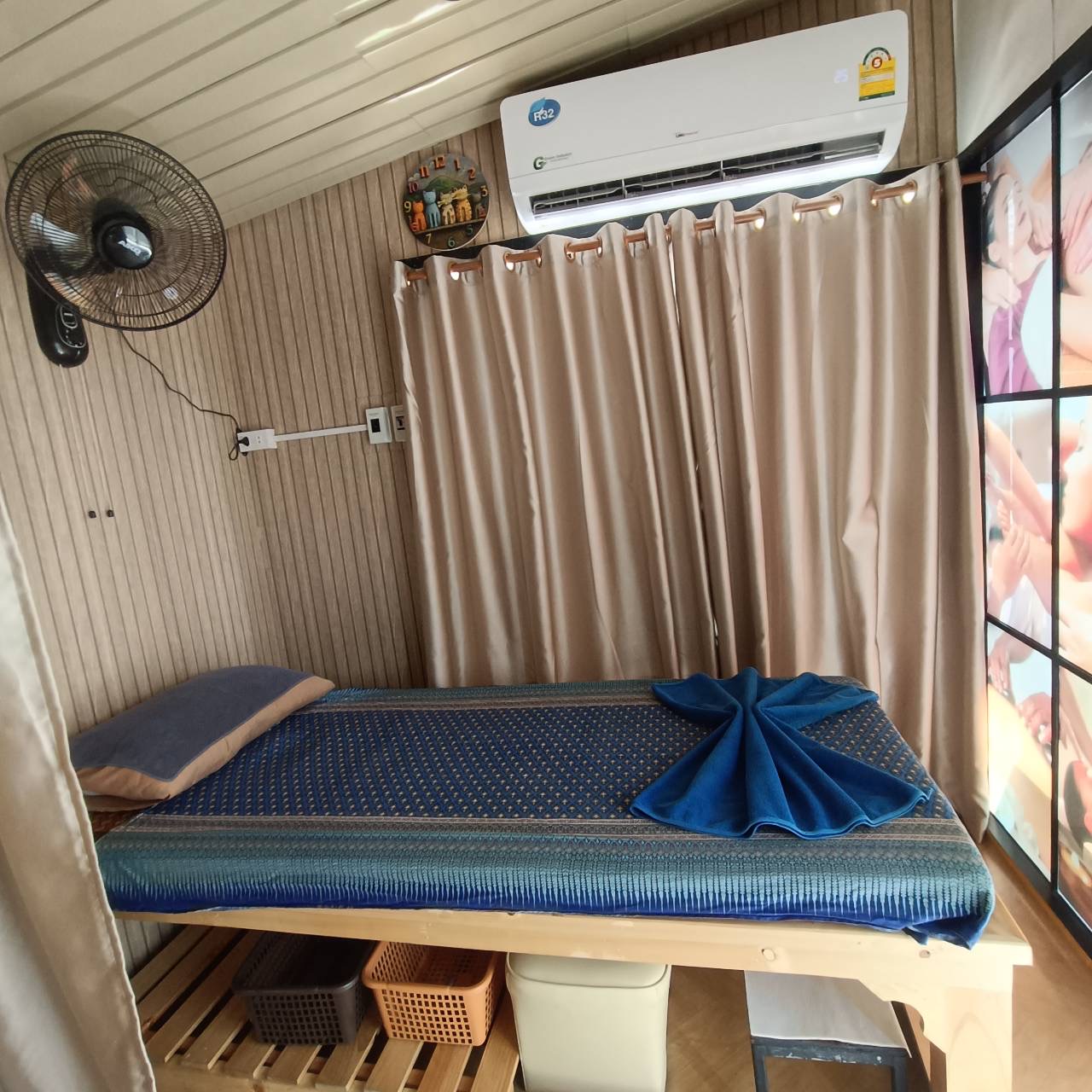 ห้องนวดแผนไทยพร้อมเตียงนวดปูผ้าลายไทยสีน้ำเงิน มีแอร์ พัดลม และผ้าม่านสีทอง