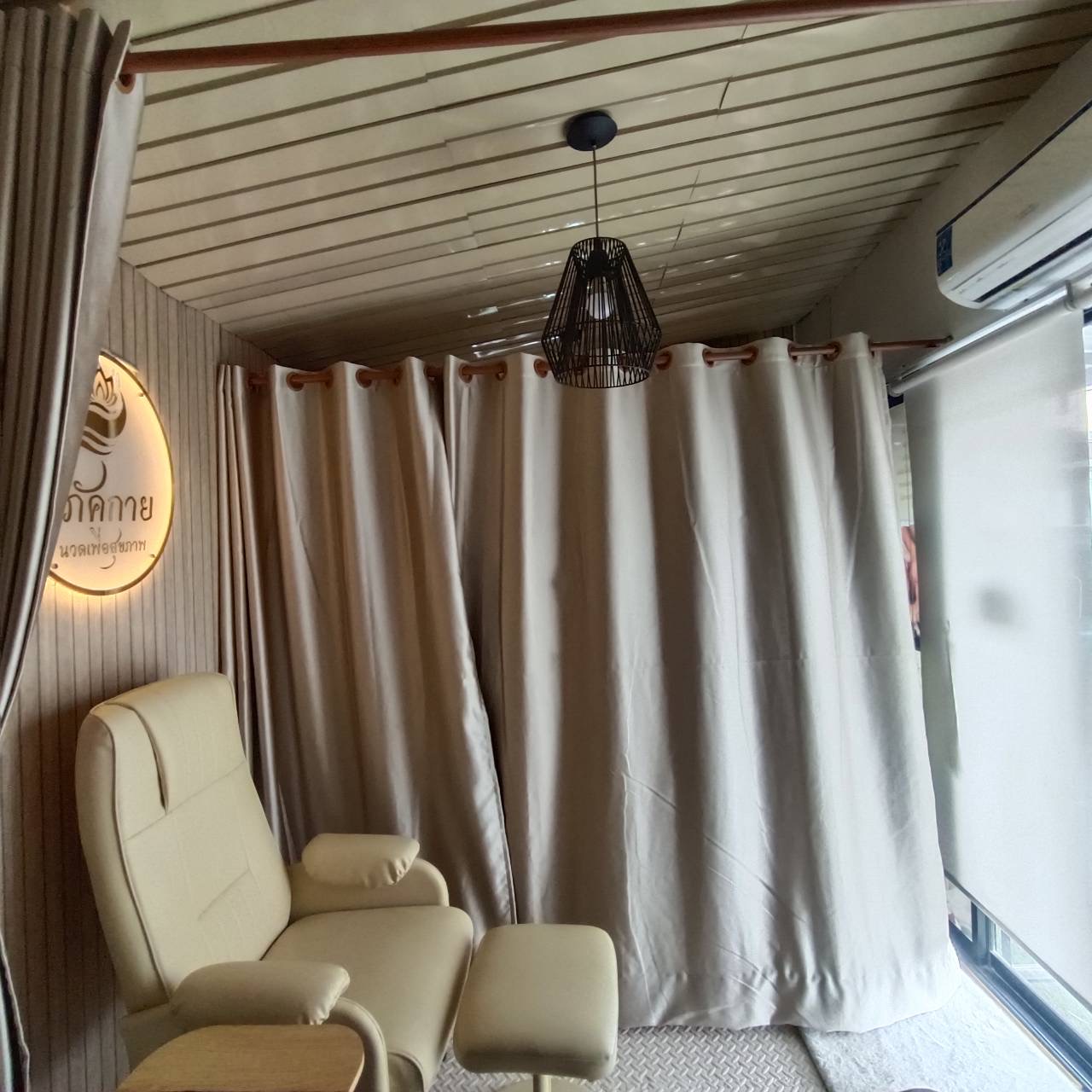 ห้องนวดสปาไทยพร้อมเก้าอี้นวดสีครีมและม่านกั้นห้อง มีป้ายไฟชื่อร้านภาคกายนวดเพื่อสุขภาพ