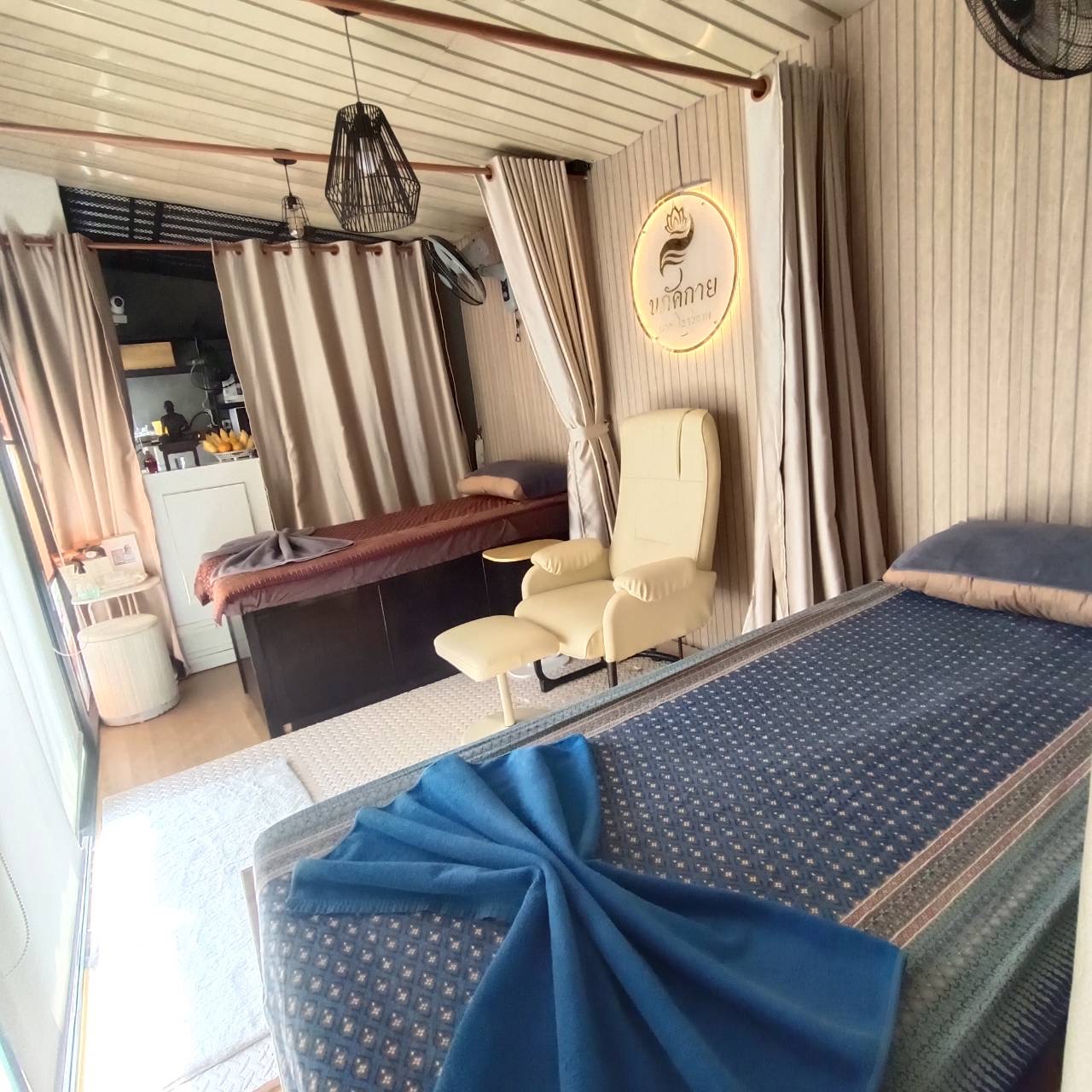 ภายในร้านนวดไทย มีเตียงนวดพร้อมผ้าปูสีน้ำเงิน เก้าอี้นวดสีครีม และม่านกั้นห้อง พร้อมป้ายชื่อร้านนกดกาย