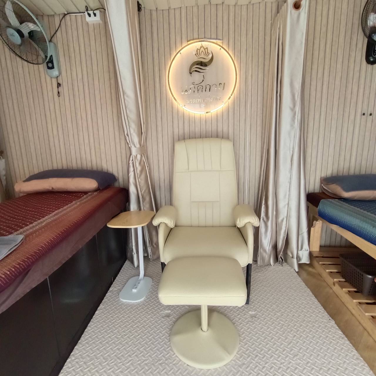 เก้าอี้นวดสีครีมพร้อมที่วางเท้าในร้านนวดไทย มีป้ายไฟโลโก้รูปดอกบัวและเตียงนวดสองข้าง