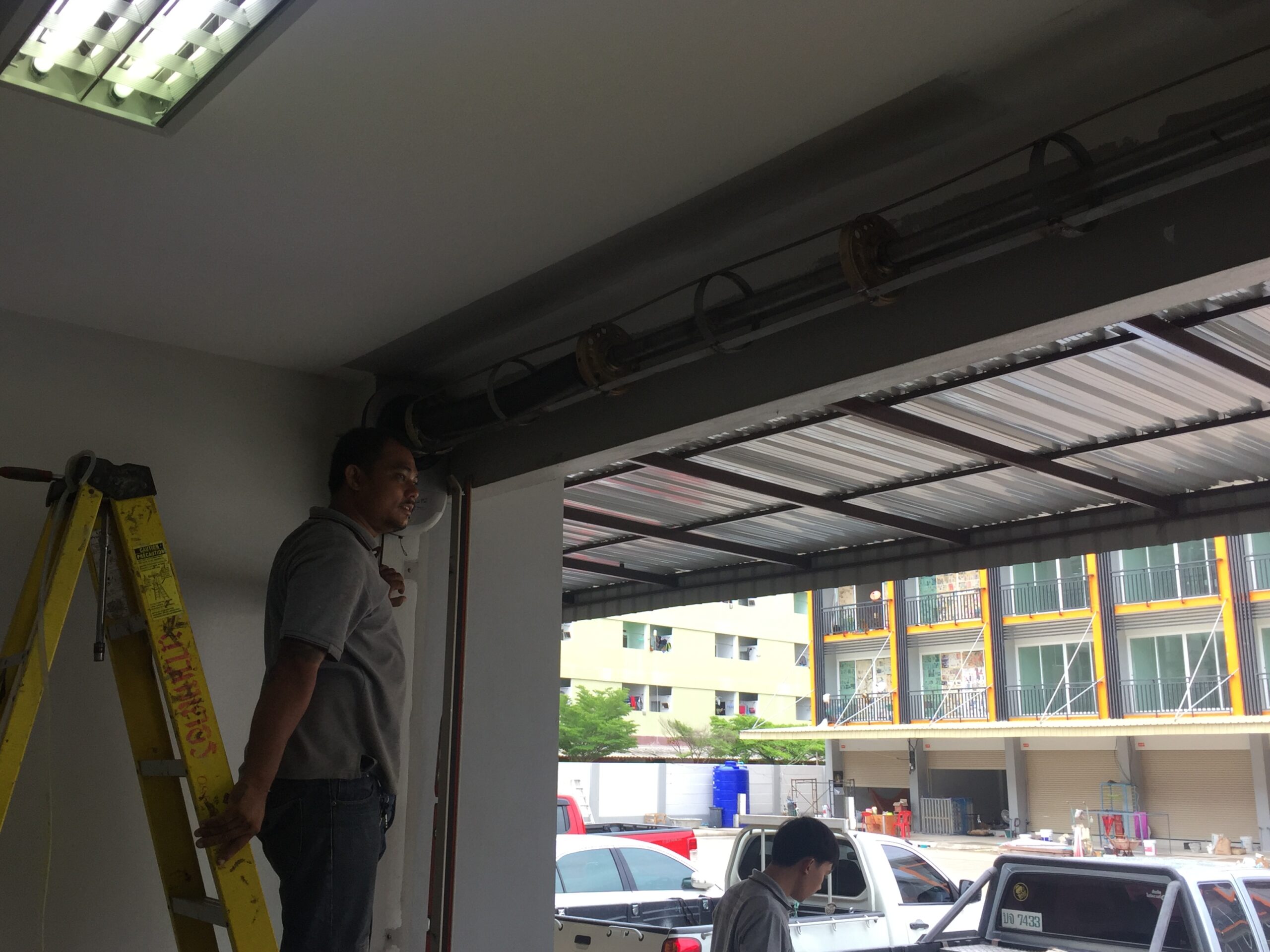 ช่างกำลังติดตั้งประตูม้วนเหล็กบริเวณหน้าร้านโดยยืนบนบันไดสีเหลือง
