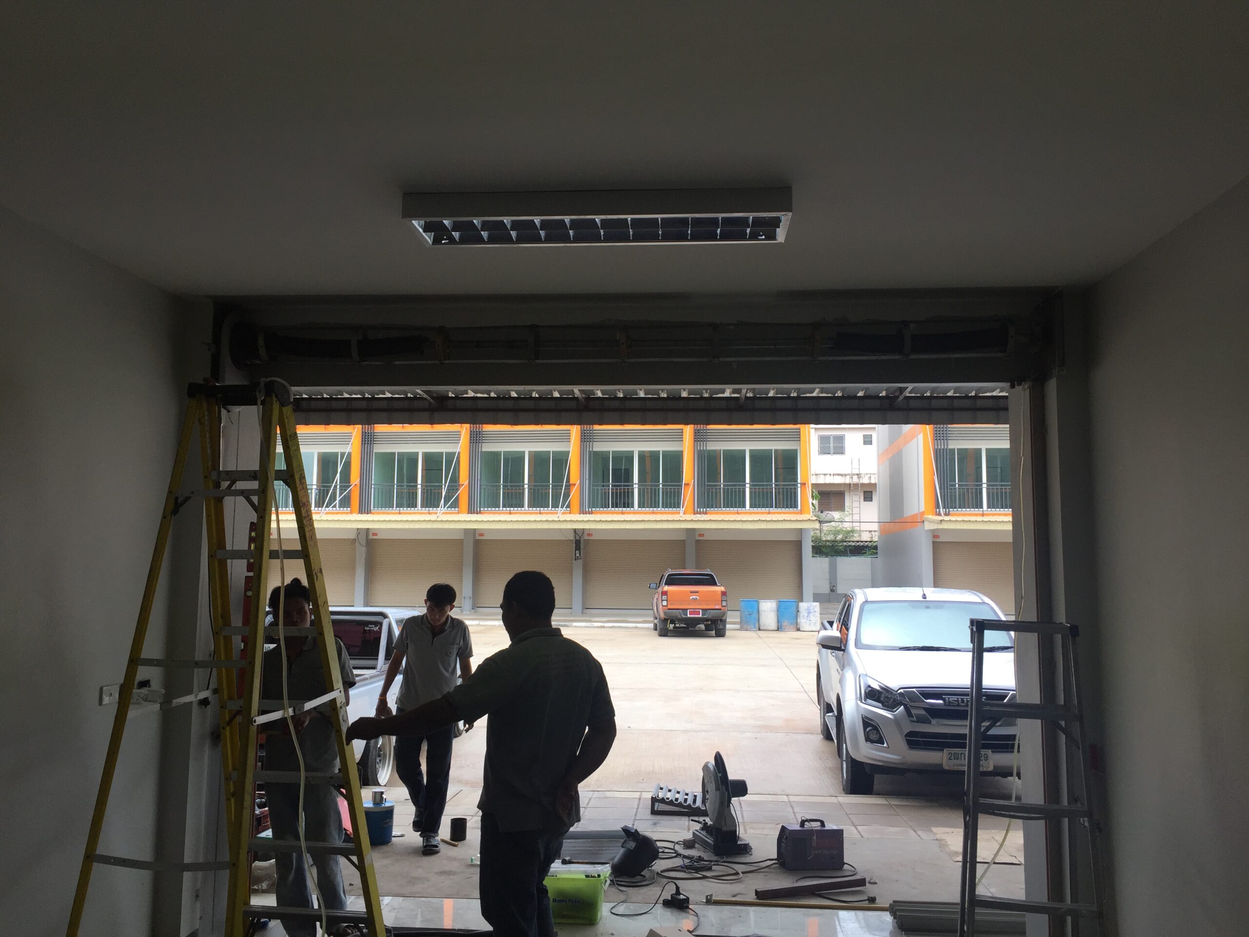 ช่างกำลังติดตั้งประตูม้วนในอาคารพาณิชย์ มีบันไดและอุปกรณ์ช่างวางอยู่