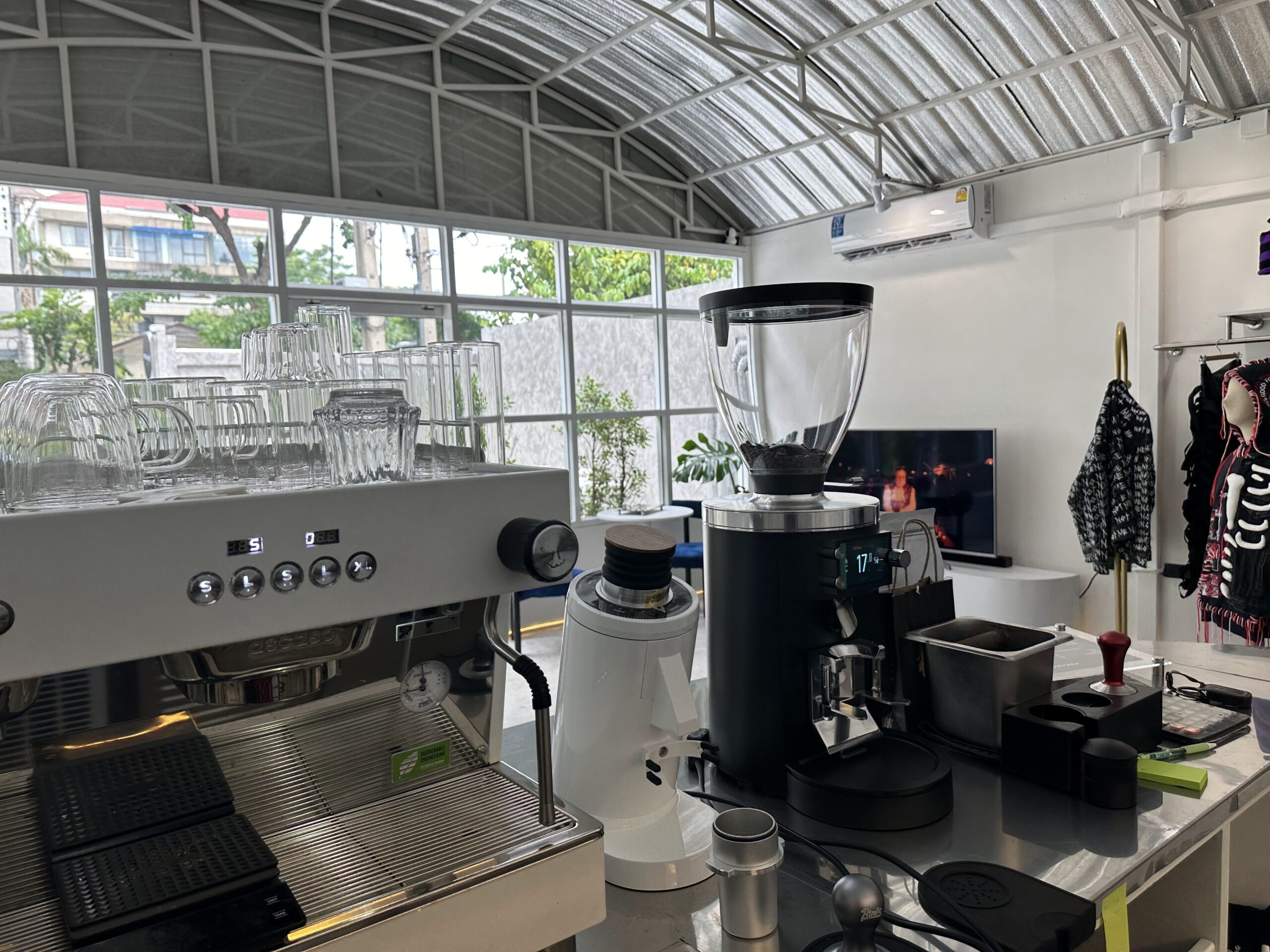 เคาน์เตอร์บาร์กาแฟพร้อมเครื่องชงเอสเปรสโซสีขาวและเครื่องบดกาแฟในร้านที่มีหลังคาโค้งและผนังกระจก