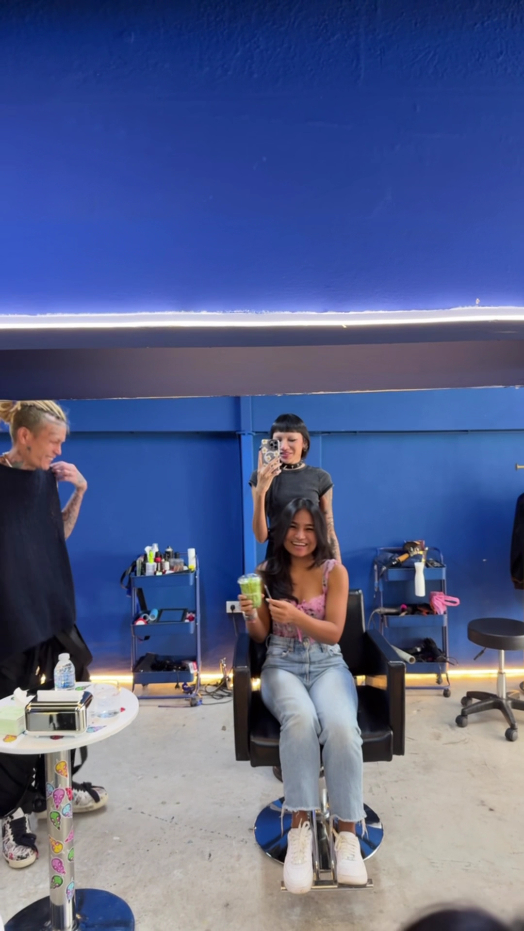 ลูกค้าสาวนั่งยิ้มบนเก้าอี้ตัดผมถือเครื่องดื่มสีเขียวในร้านทำผมผนังสีน้ำเงินขณะช่างผมถ่ายรูปผ่านกระจก