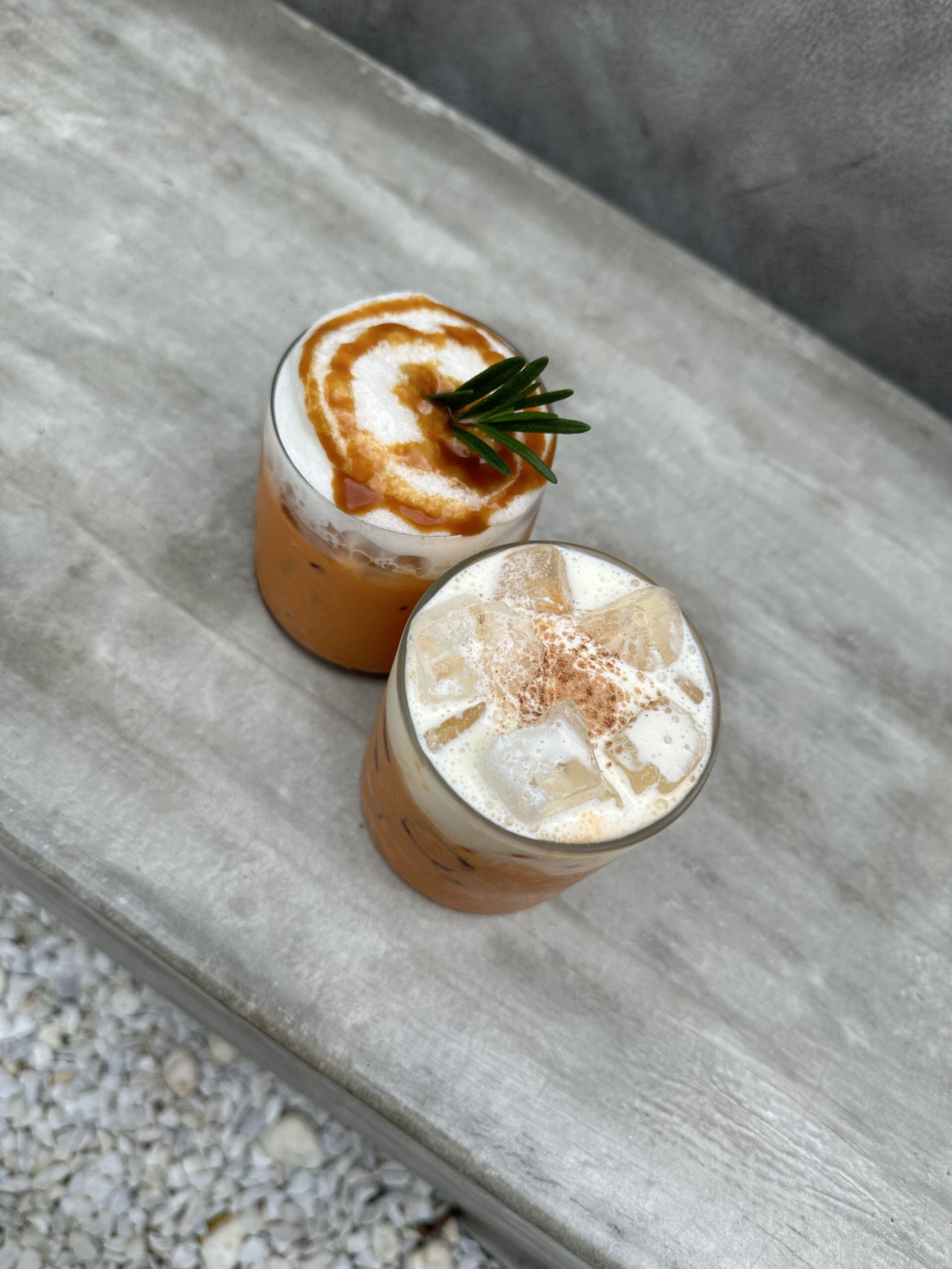 เครื่องดื่มชาไทยเย็นสองแก้วบนโต๊ะปูน แก้วแรกมีฟองนมและซอสคาราเมลตกแต่งด้วยใบโรสแมรี่ แก้วที่สองเป็นชาไทยเย็นใส่น้ำแข็งโรยผงอบเชย