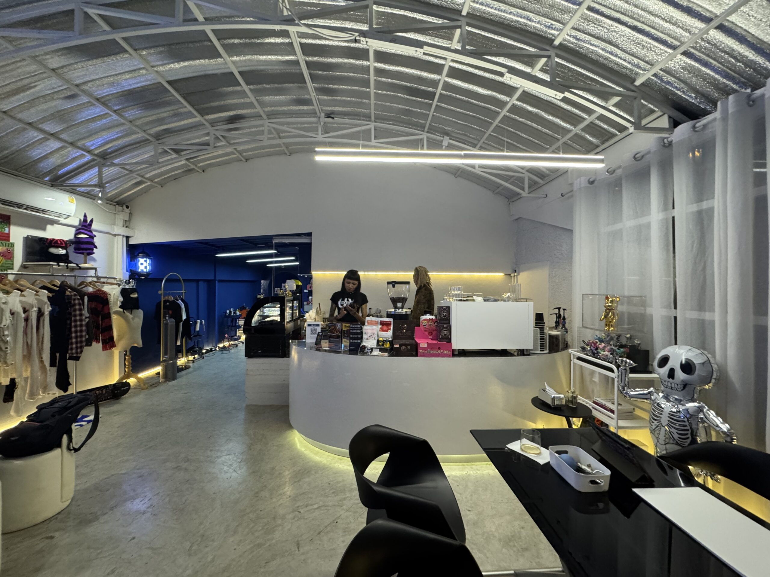 ร้านกาแฟสไตล์อินดัสเทรียลพร้อมเคาน์เตอร์สีขาวโค้ง เครื่องชงกาแฟ ราวแขวนเสื้อผ้า และตุ๊กตาโครงกระดูกสีเงินตกแต่ง ภายใต้หลังคาโค้งโครงเหล็ก