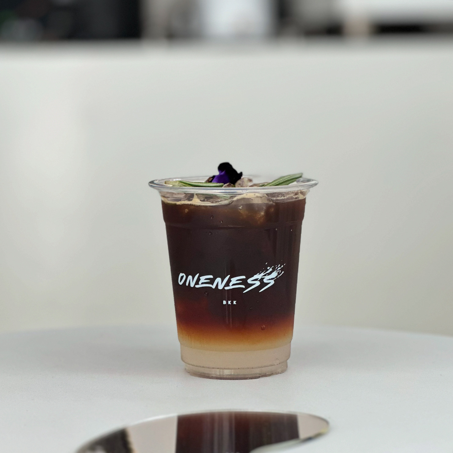 เครื่องดื่มกาแฟเย็นแบบเลเยอร์ในแก้วพลาสติกโลโก้ Oneness BKK ตกแต่งด้วยดอกอัญชันและใบโรสแมรี่ วางบนโต๊ะสีขาว