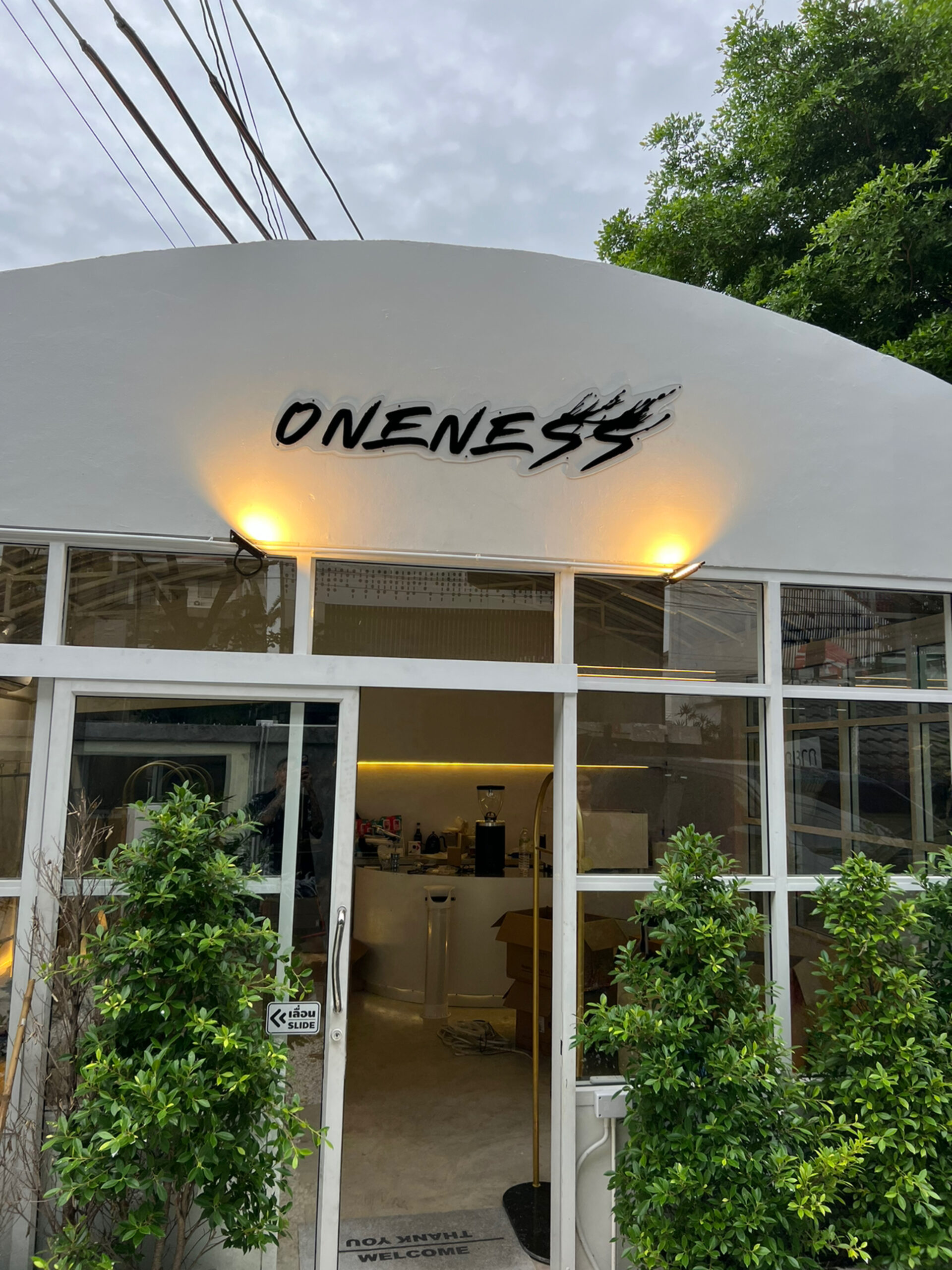ด้านหน้าร้าน ONENESS คาเฟ่สีขาวสไตล์โมเดิร์นพร้อมป้ายชื่อร้านสีดำ ประตูกระจกบานเลื่อน และต้นไม้สีเขียวตกแต่งด้านหน้า