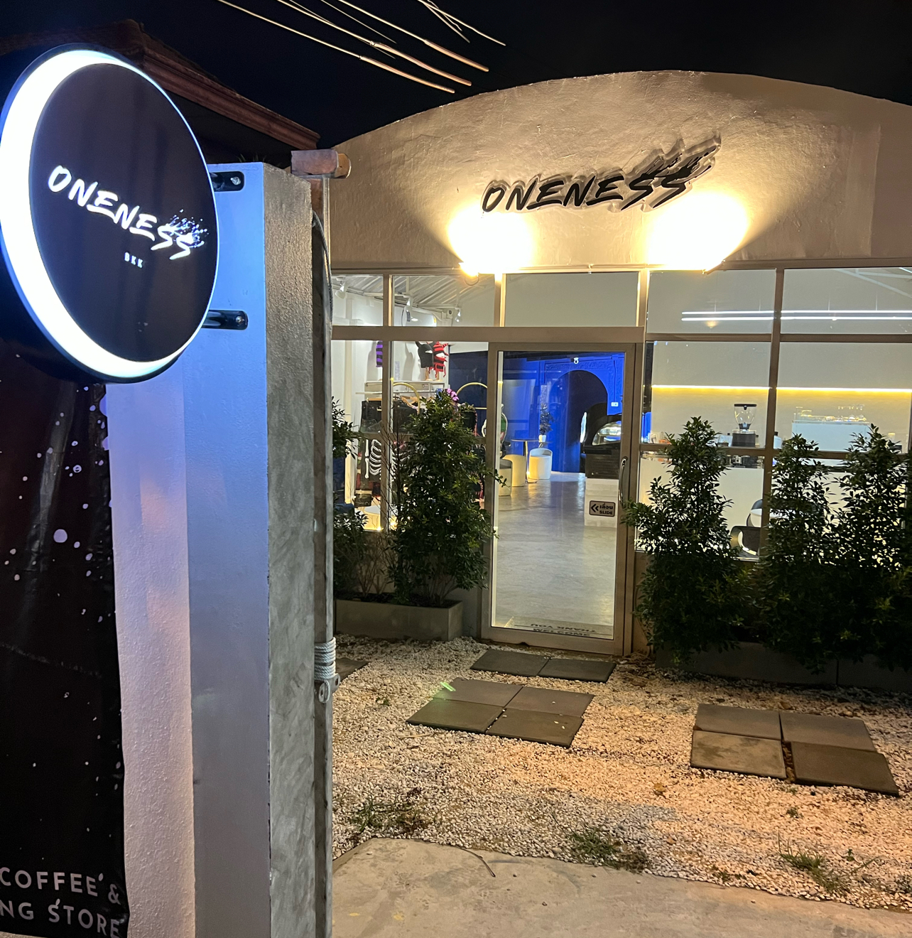 ทางเข้าร้าน Oneness Coffee & Store ในเวลากลางคืน มีป้ายไฟวงกลมส่องสว่าง ต้นไม้เขียวขนาบสองข้าง และด้านในร้านตกแต่งสวยงาม