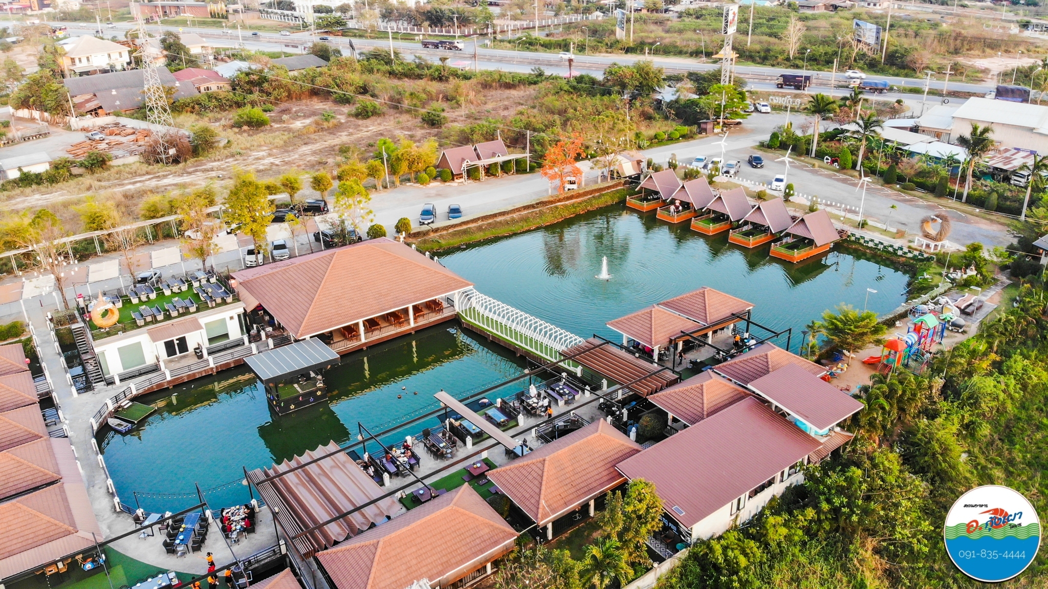 ภาพถ่ายมุมสูงของร้านอาหารริมน้ำพร้อมศาลาลอยน้ำ สระว่ายน้ำ และน้ำพุกลางบ่อ บรรยากาศสวยงาม