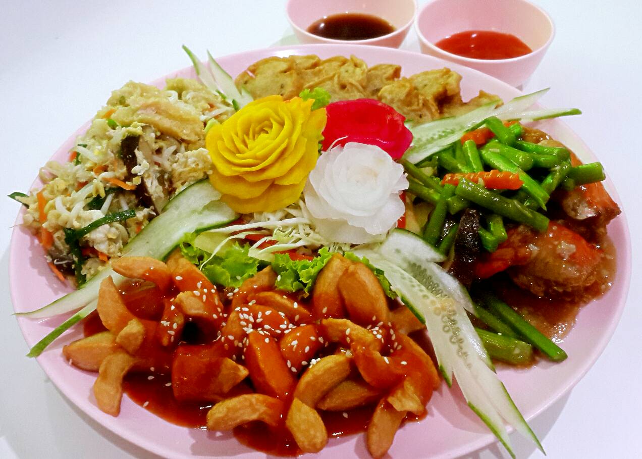 จานอาหารจีนรวมมิตรบนจานสีชมพู มีผัดผักรวม ไก่ผัดซอส เกี๊ยวทอด และผักสดตกแต่งด้วยดอกไม้แกะสลัก พร้อมน้ำจิ้ม