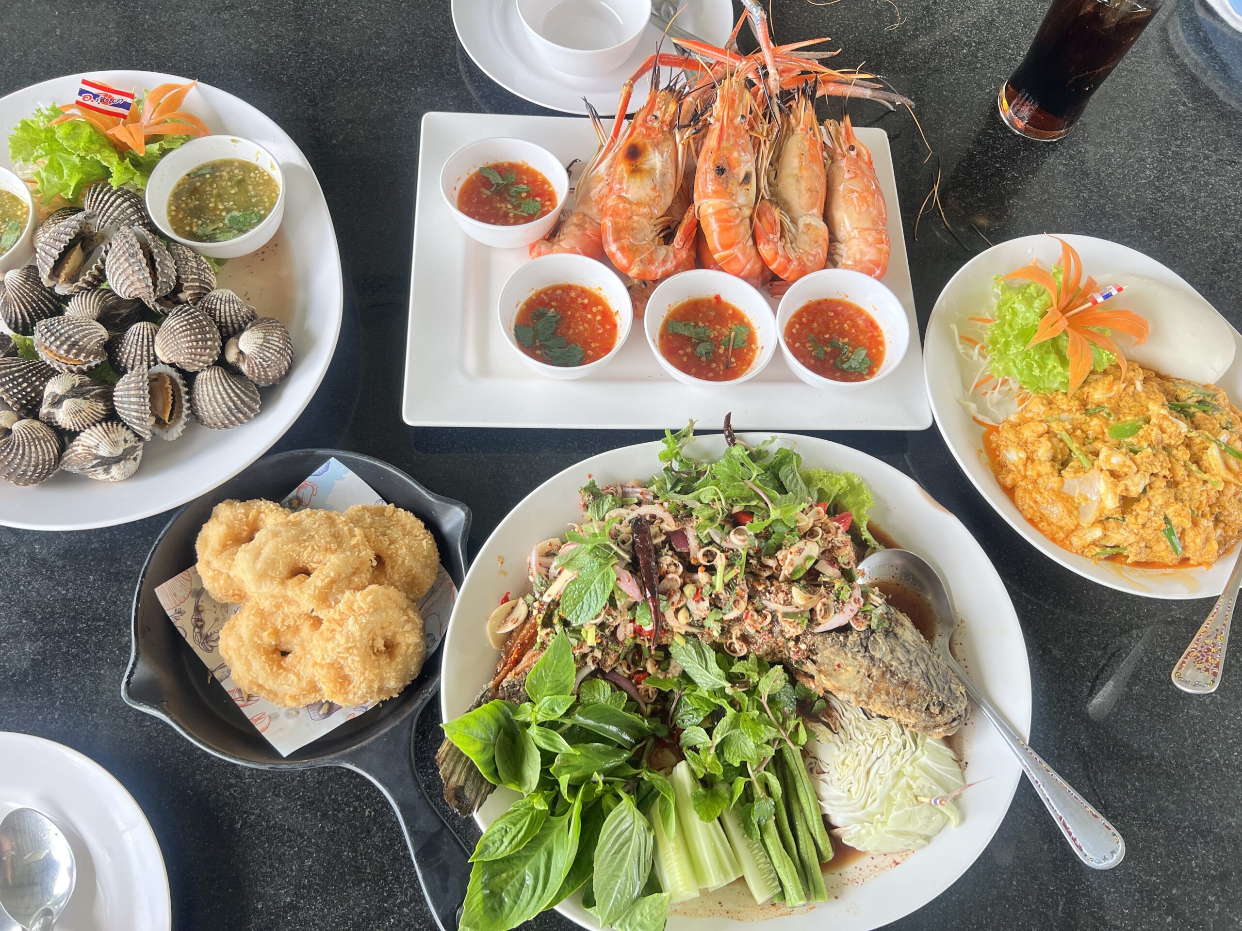 สำรับอาหารทะเลไทยบนโต๊ะ มีกุ้งแม่น้ำเผา หอยแครงลวก ปลาสมุนไพรทอดราดยำ ไข่เจียว และขนมทอดกรอบ พร้อมน้ำจิ้มซีฟู้ด