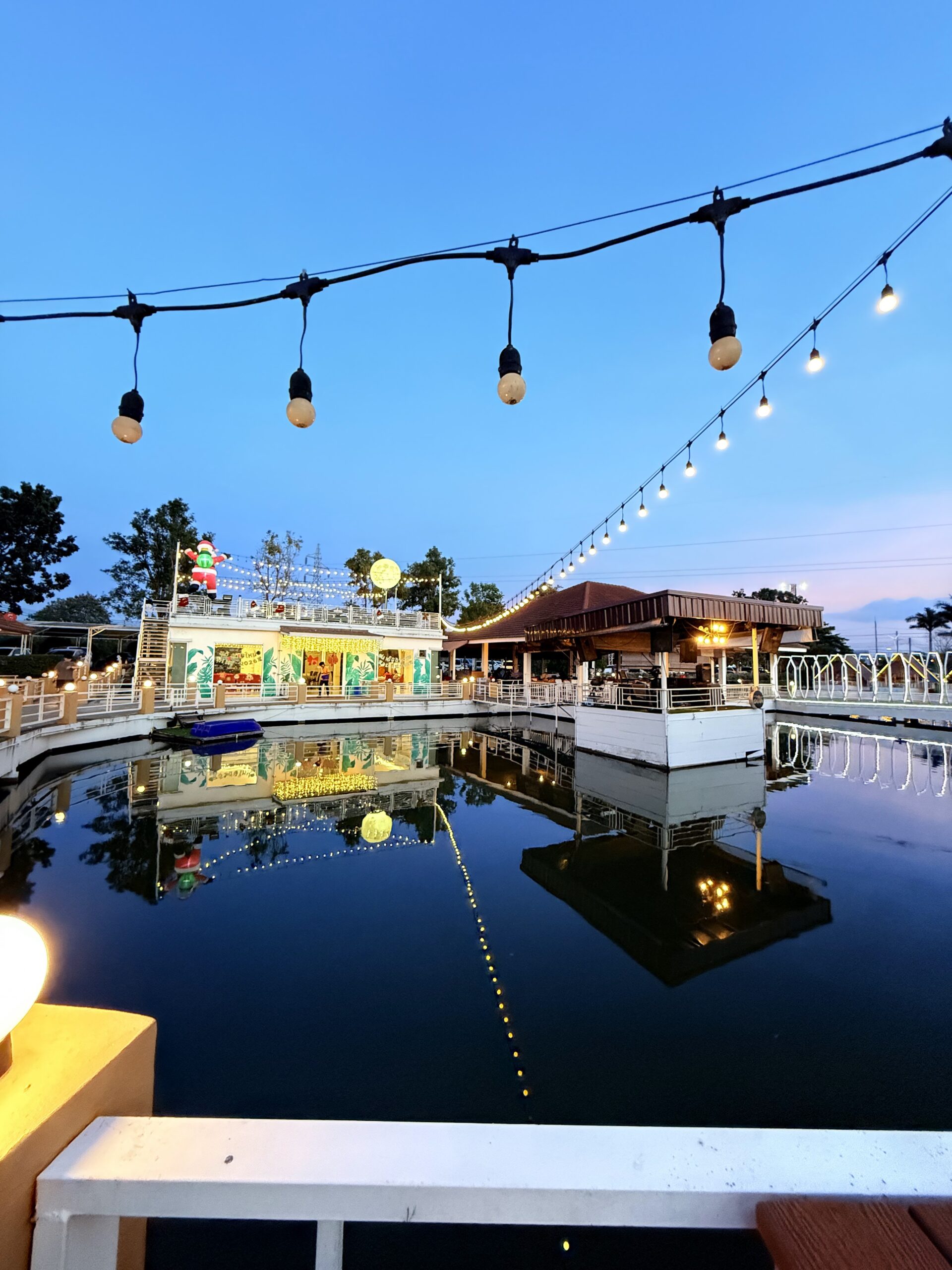 ร้านอาหารริมน้ำยามพลบค่ำ ประดับไฟสายสวยงาม มีสะพานทางเดินสีขาวและอาคารตกแต่งสีสันสดใส สะท้อนเงาลงในผืนน้ำ