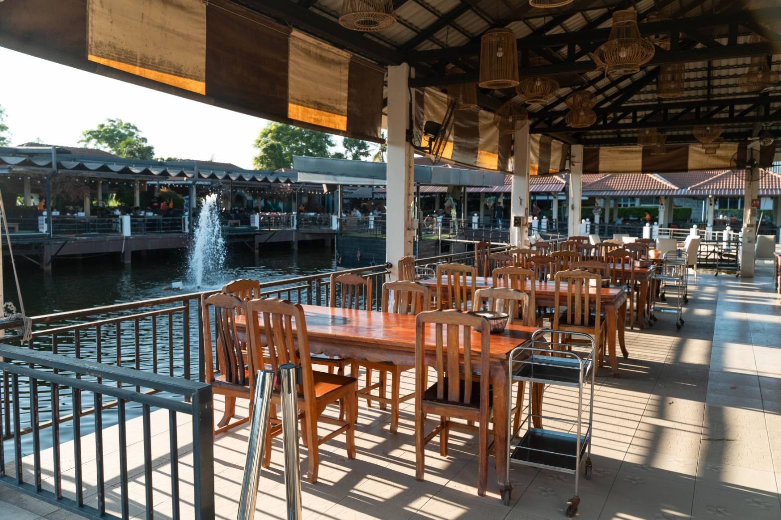 ร้านอาหารริมน้ำกลางแจ้งพร้อมโต๊ะและเก้าอี้ไม้เรียงรายริมรั้วกั้นน้ำ มีน้ำพุอยู่กลางสระ บรรยากาศร่มรื่นยามบ่าย