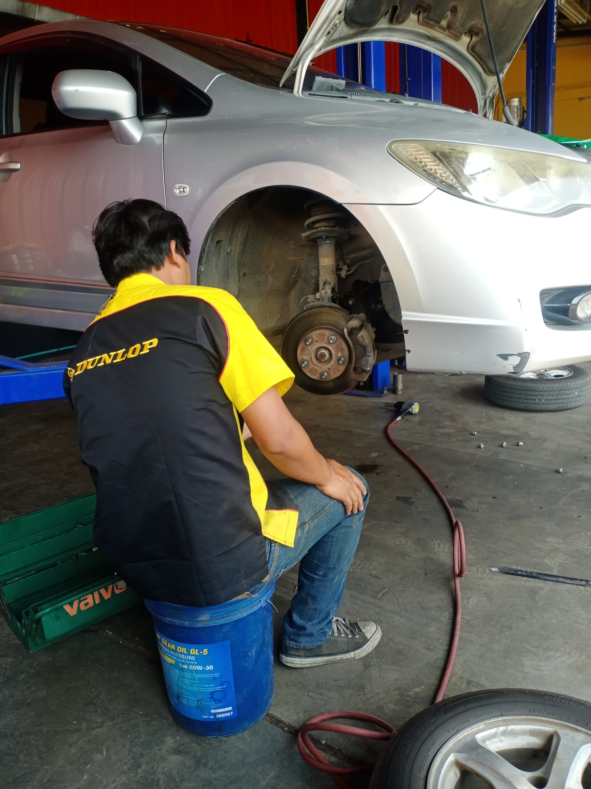 ช่างซ่อมรถสวมเสื้อ Dunlop นั่งบนถังน้ำมันเกียร์ตรวจสอบระบบเบรกรถยนต์สีเงินในอู่ซ่อมรถ