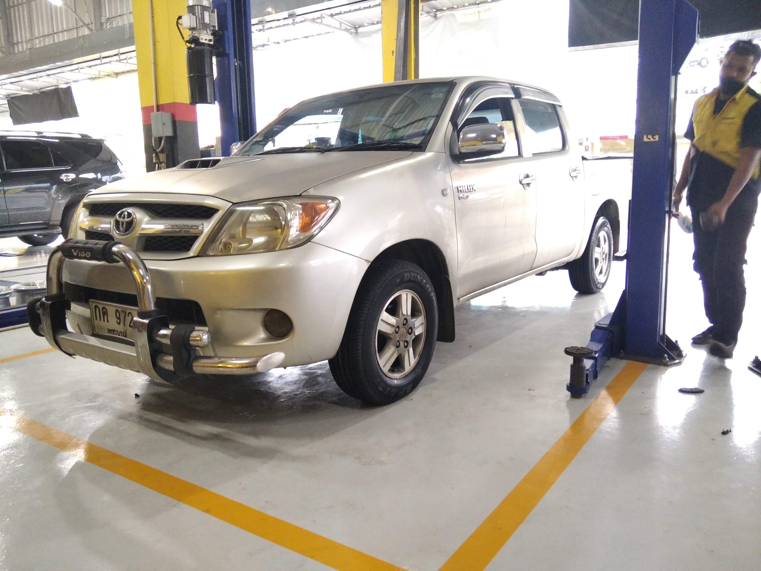 รถกระบะโตโยต้า ไฮลักซ์ วีโก้ สีเงิน จอดอยู่ในศูนย์บริการซ่อมรถยนต์ มีช่างยืนอยู่ข้างๆ