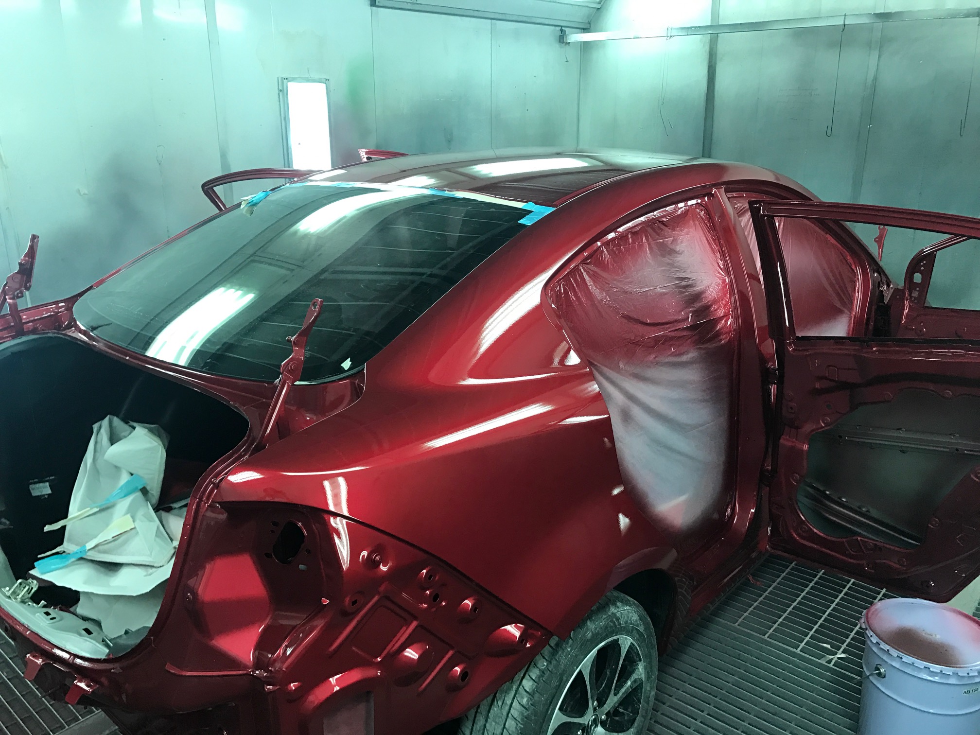 รถยนต์สีแดงกำลังถูกพ่นสีในห้องพ่นสี โดยมีการถอดชิ้นส่วนและปิดมาสกิ้งเทปป้องกัน