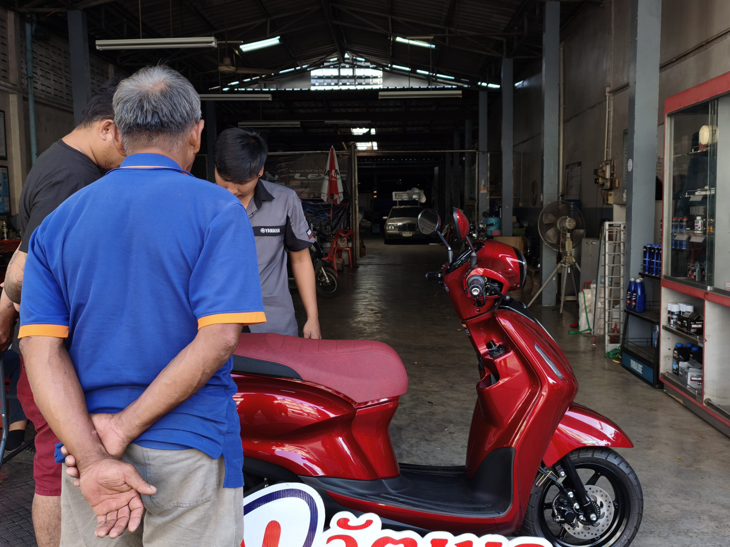 ลูกค้ากำลังดูรถมอเตอร์ไซค์สกู๊ตเตอร์สีแดงเงาในร้านซ่อมรถ มีพนักงานยามาฮ่าให้บริการ