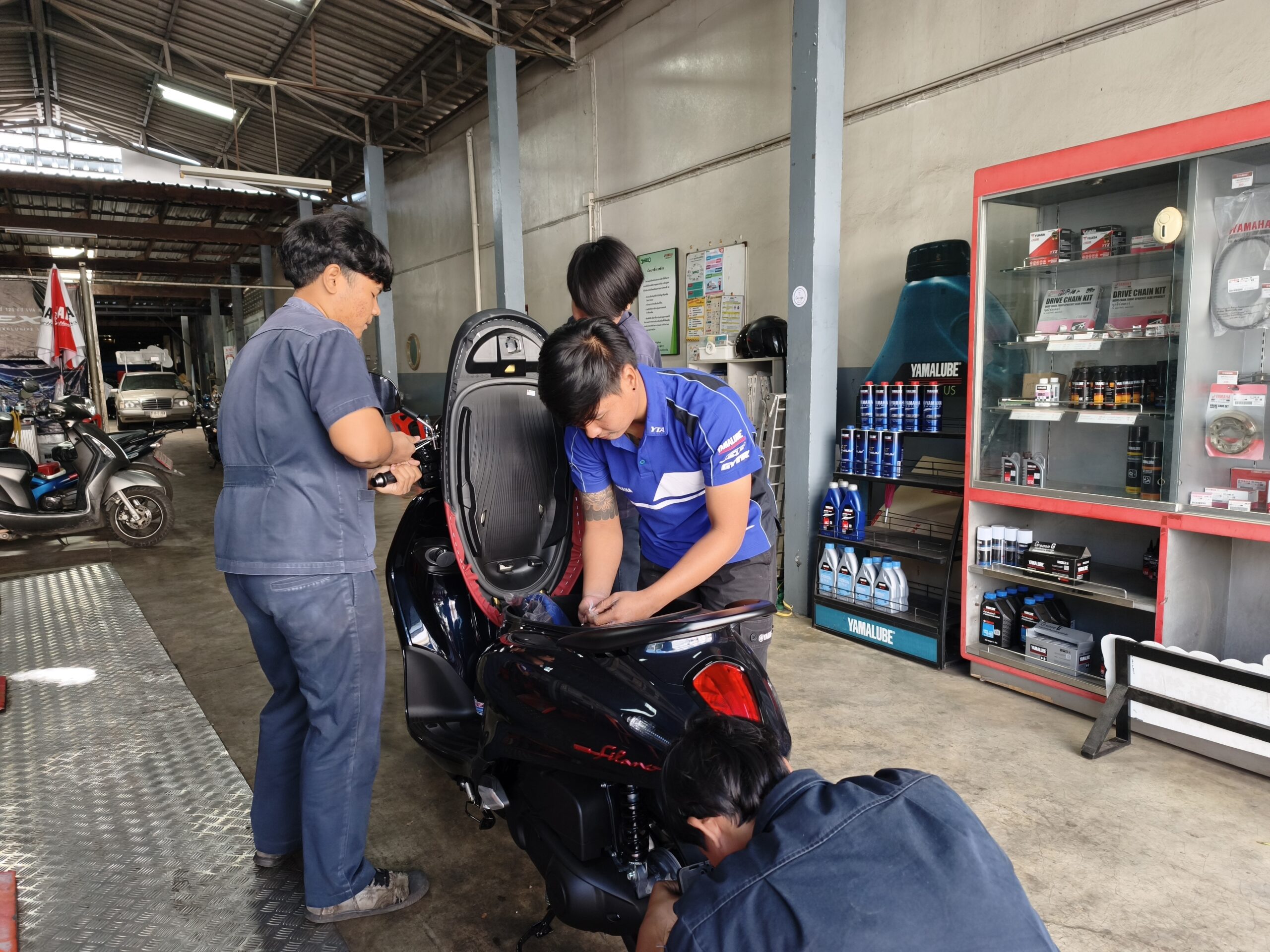 ช่างซ่อมมอเตอร์ไซค์ยามาฮ่ากำลังตรวจสอบรถสกู๊ตเตอร์สีดำในศูนย์บริการ พร้อมผลิตภัณฑ์ยามาลูปจัดแสดงในตู้กระจก
