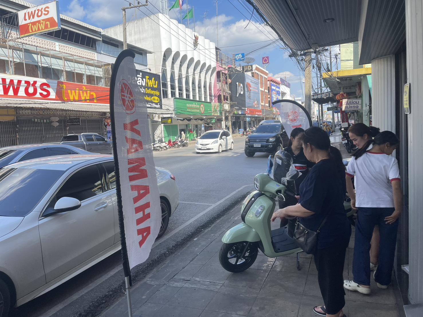 ถนนในเมืองไทยที่มีธงโฆษณายามาฮ่าและรถมอเตอร์ไซค์สีเขียวอ่อนจอดอยู่บนทางเท้า มีผู้คนยืนดูอยู่หน้าร้าน