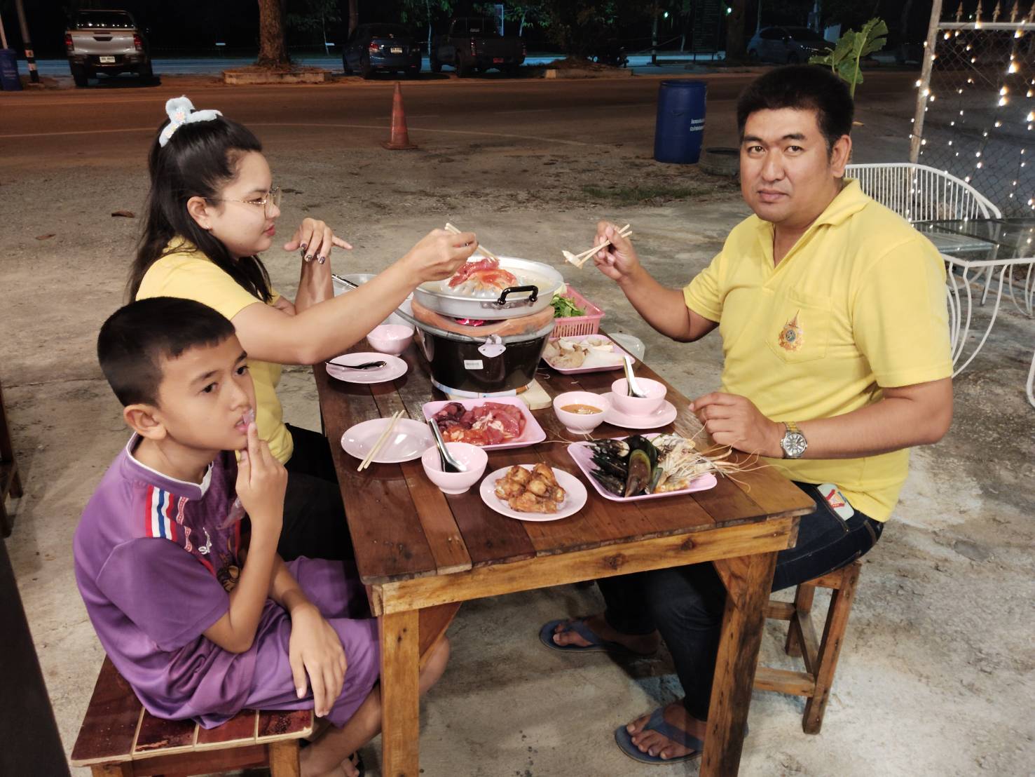 ครอบครัวนั่งทานหมูกระทะและอาหารทะเลที่ร้านอาหารกลางแจ้งในเวลากลางคืน มีพ่อแม่ลูกใช้ตะเกียบคีบเนื้อย่างบนเตา
