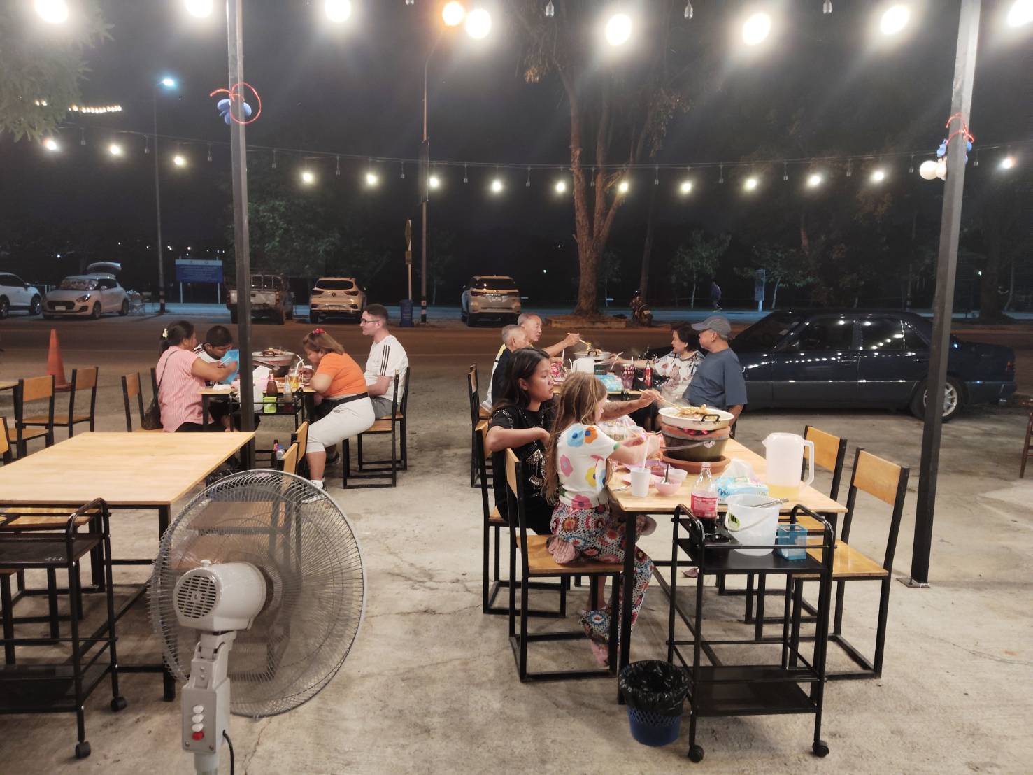 ร้านอาหารกลางแจ้งตอนกลางคืนมีลูกค้านั่งทานอาหารหลายโต๊ะ ตกแต่งด้วยไฟสายประดับ มีพัดลมตั้งพื้นและที่จอดรถด้านหลัง