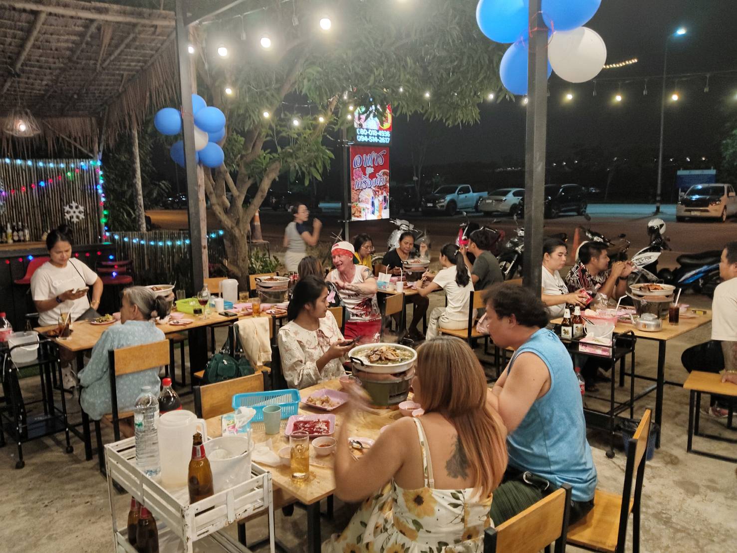 บรรยากาศร้านอาหารกลางแจ้งยามค่ำคืน ลูกค้านั่งทานอาหารหลายโต๊ะ มีลูกโป่งสีฟ้าขาวตกแต่ง ไฟสายประดับ และหม้อไฟบนโต๊ะ