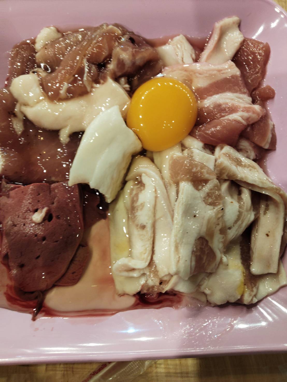 จานเนื้อสดรวมมิตรพร้อมไข่แดงสำหรับปิ้งย่าง มีหมูสามชั้น ตับ และเนื้อหมักซอส บนจานสีชมพู