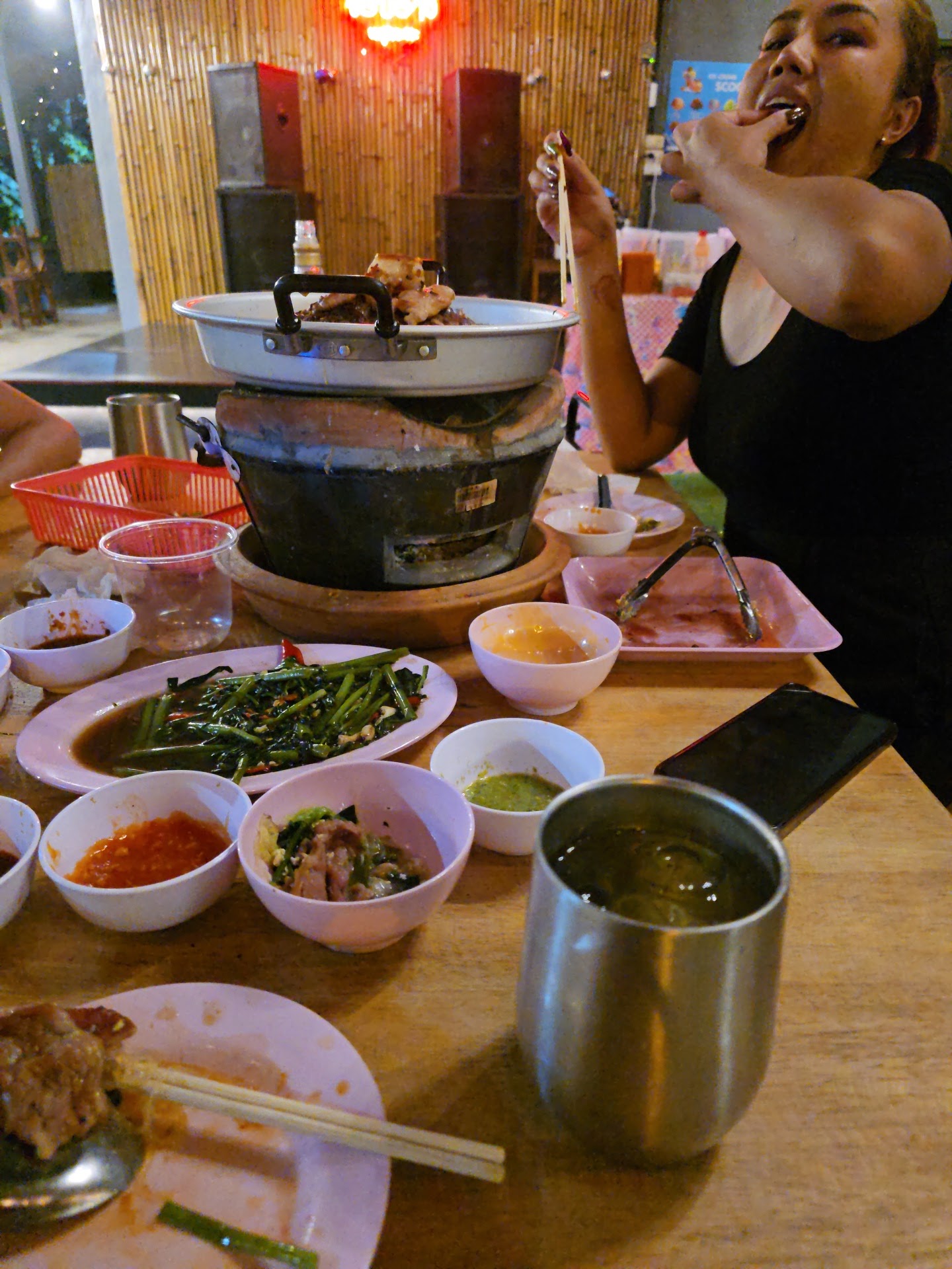 ผู้หญิงกำลังรับประทานหมูกระทะพร้อมเครื่องเคียงหลากหลาย น้ำจิ้มหลายชนิด ผัดผักบุ้ง และเตาปิ้งย่างบนโต๊ะไม้ในร้านอาหารบรรยากาศสบายๆ