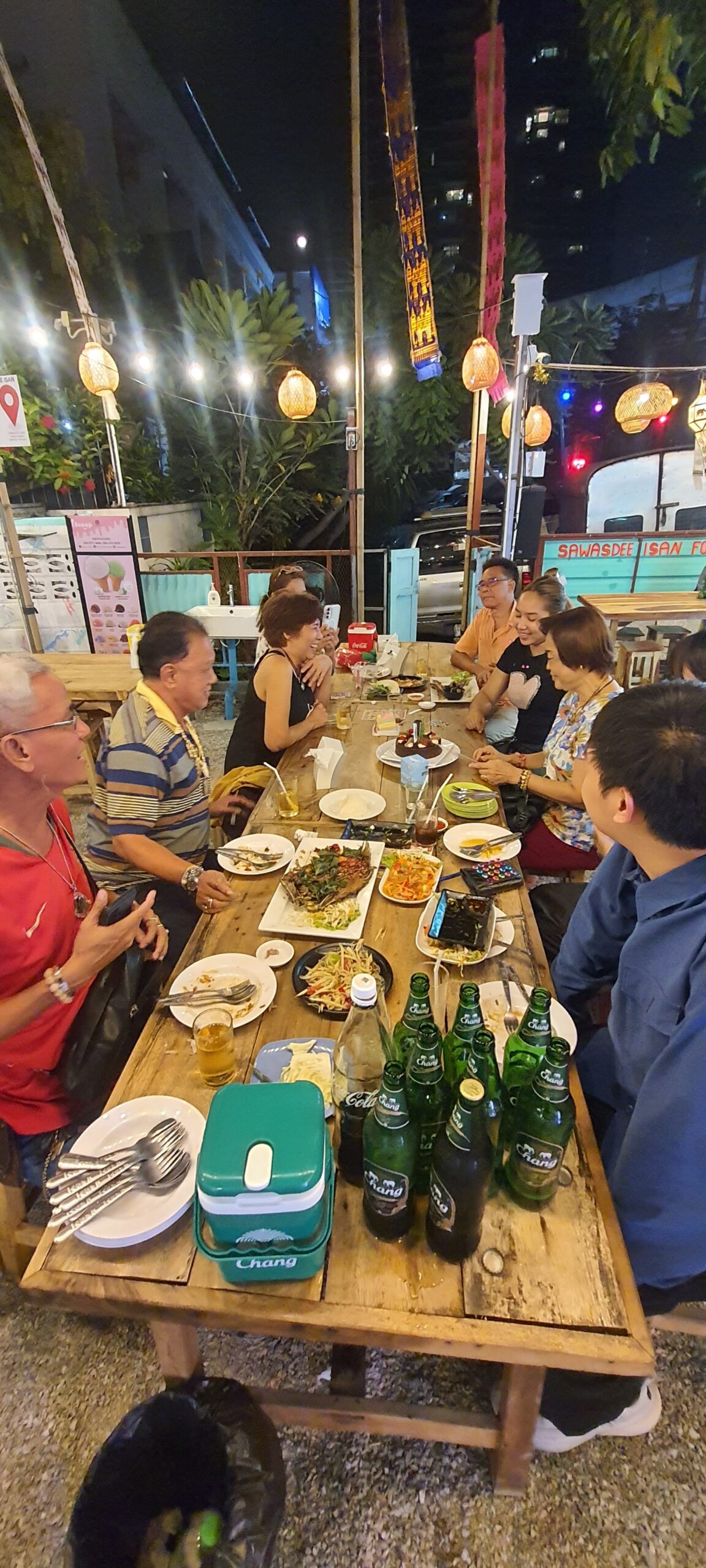 กลุ่มเพื่อนนั่งรับประทานอาหารอีสานที่ร้านสวัสดีอีสานฟู้ด พร้อมเบียร์ช้างและอาหารไทยบนโต๊ะไม้กลางแจ้งยามค่ำคืน