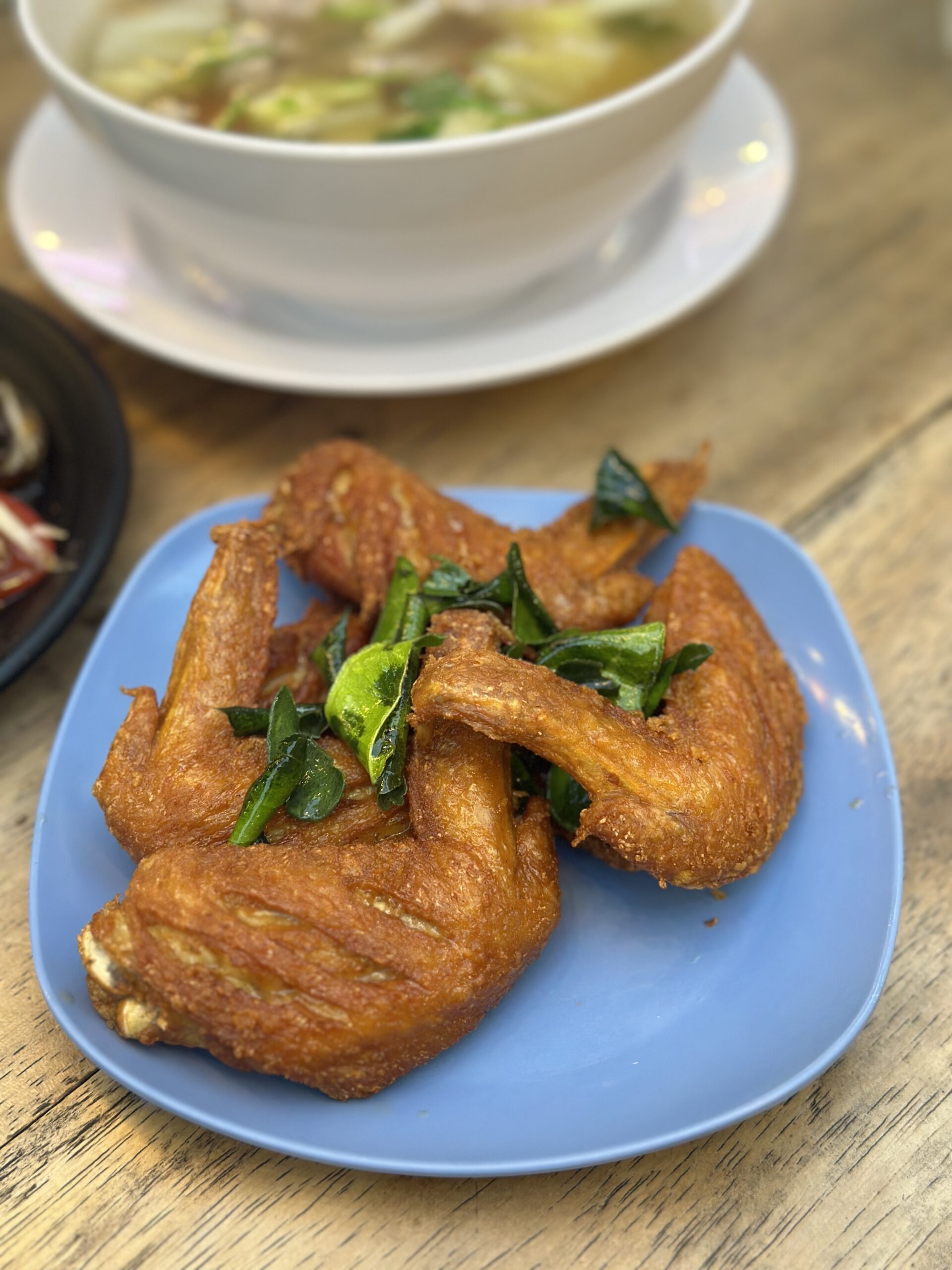 ปีกไก่ทอดกรอบโรยใบโหระพา เสิร์ฟบนจานสีฟ้า พร้อมชามต้มจืดอยู่ด้านหลัง