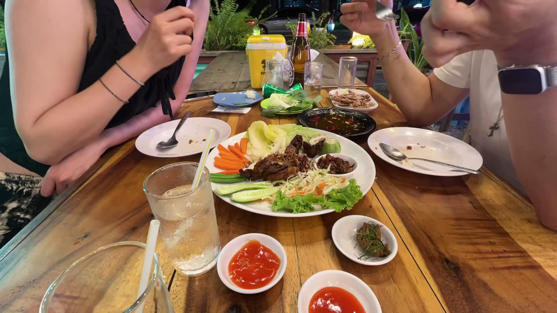 จานเนื้อย่างพร้อมผักสดและน้ำจิ้มแดงบนโต๊ะไม้ในร้านอาหารกลางแจ้ง มีเบียร์และเครื่องดื่มเย็น