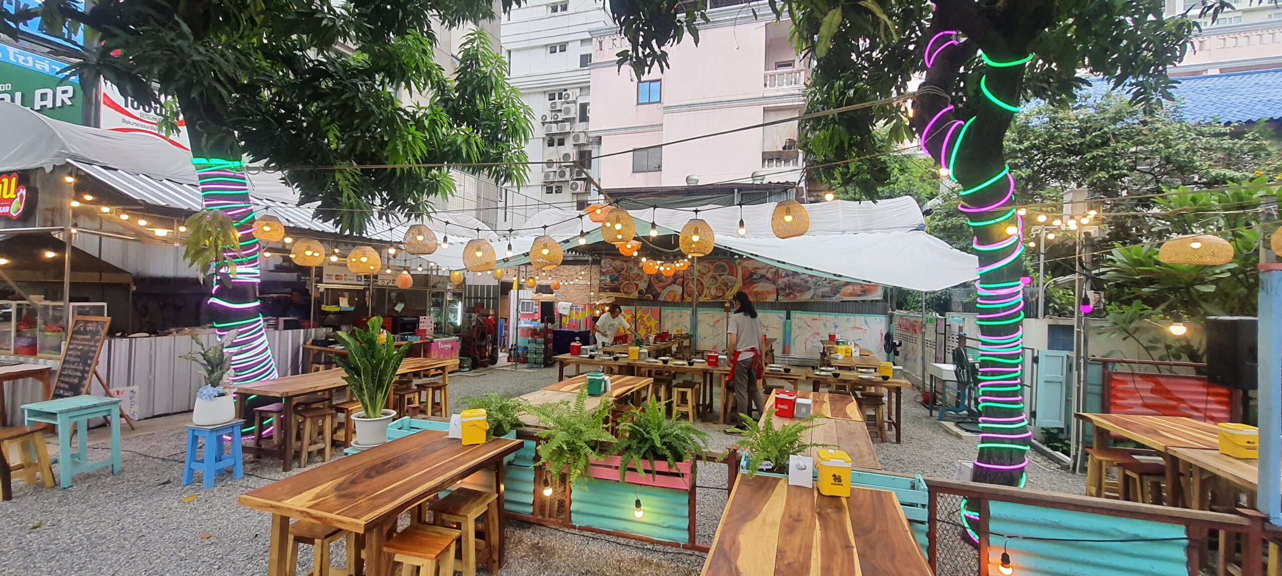 ร้านอาหารกลางแจ้งตกแต่งด้วยไฟนีออนสีเขียวและชมพูพันรอบต้นไม้ โคมไฟหวายแขวนประดับ โต๊ะไม้เก้าอี้ไม้จัดเรียงในลานกรวด บรรยากาศสบายๆ สไตล์สตรีทฟู้ด