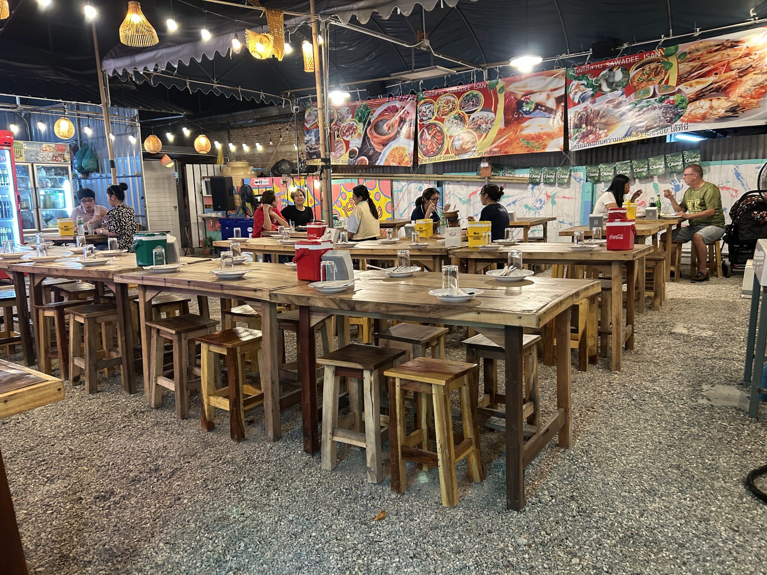 บรรยากาศร้านสวัสดีอีสานในยามค่ำคืน มีโต๊ะไม้และเก้าอี้ไม้จัดเรียงกลางแจ้ง ป้ายเมนูอาหารขนาดใหญ่และป้ายเบียร์ช้างตกแต่งผนัง มีลูกค้านั่งรับประทานอาหาร