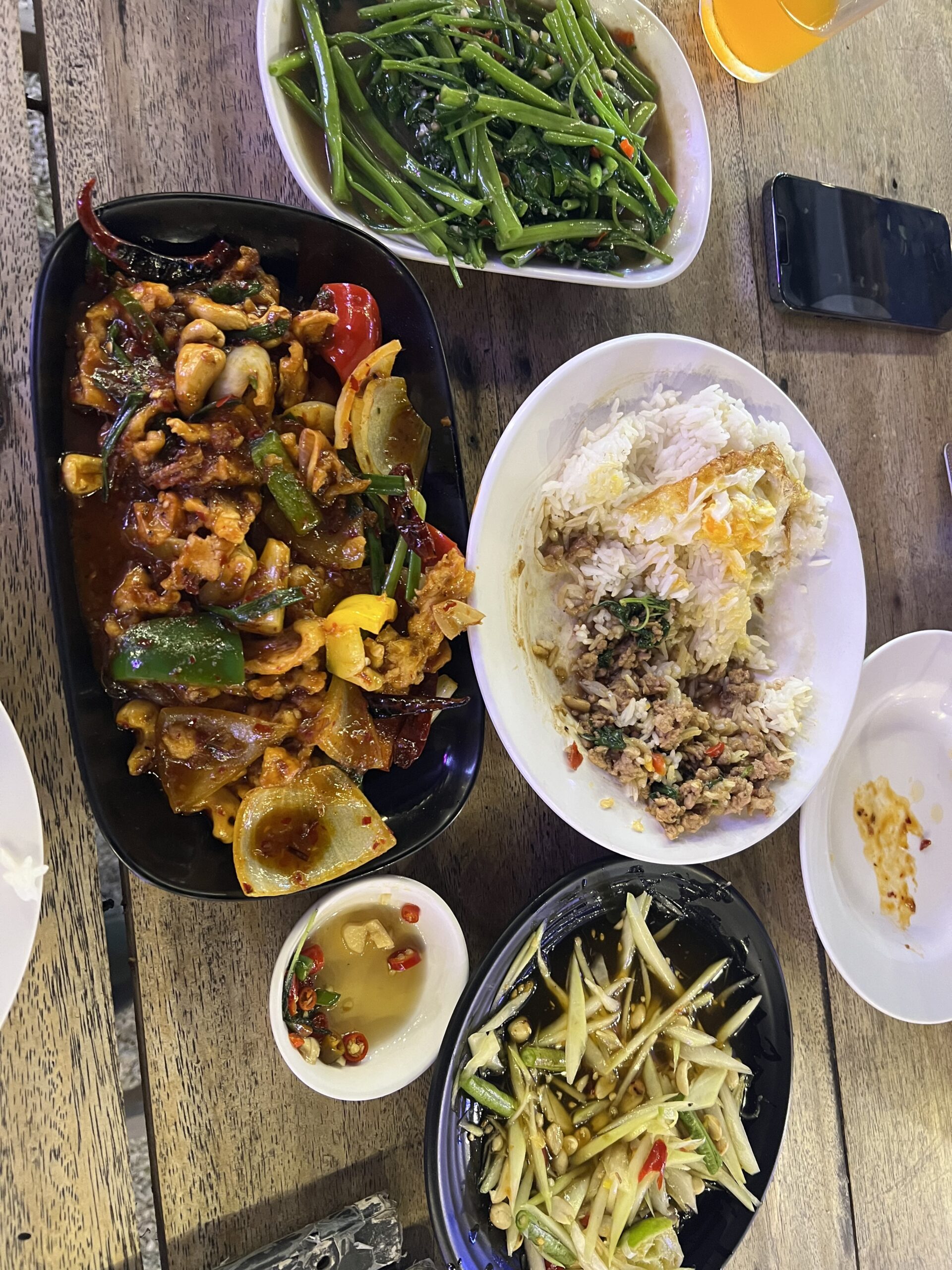 อาหารไทยหลายจานบนโต๊ะไม้ ประกอบด้วยผัดเผ็ดไก่กับเม็ดมะม่วงหิมพานต์ ผัดผักบุ้ง ข้าวราดกะเพราหมูสับไข่ดาว ส้มตำมะม่วง และน้ำปลาพริก