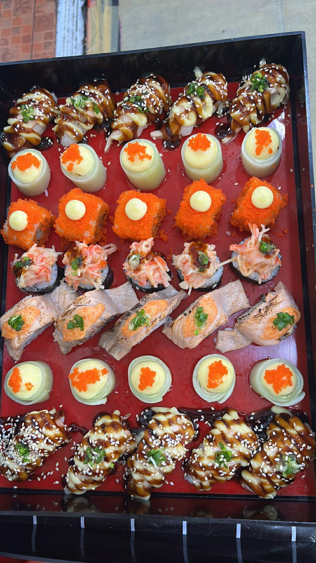 จานซูชิรวมหลากหลายชนิดบนถาดสีแดง มีซูชิราดซอสเทอริยากิโรยงา ซูชิไข่ปลาบิน ซูชิแซลมอนย่างไฟ และกุนกันครีมชีส